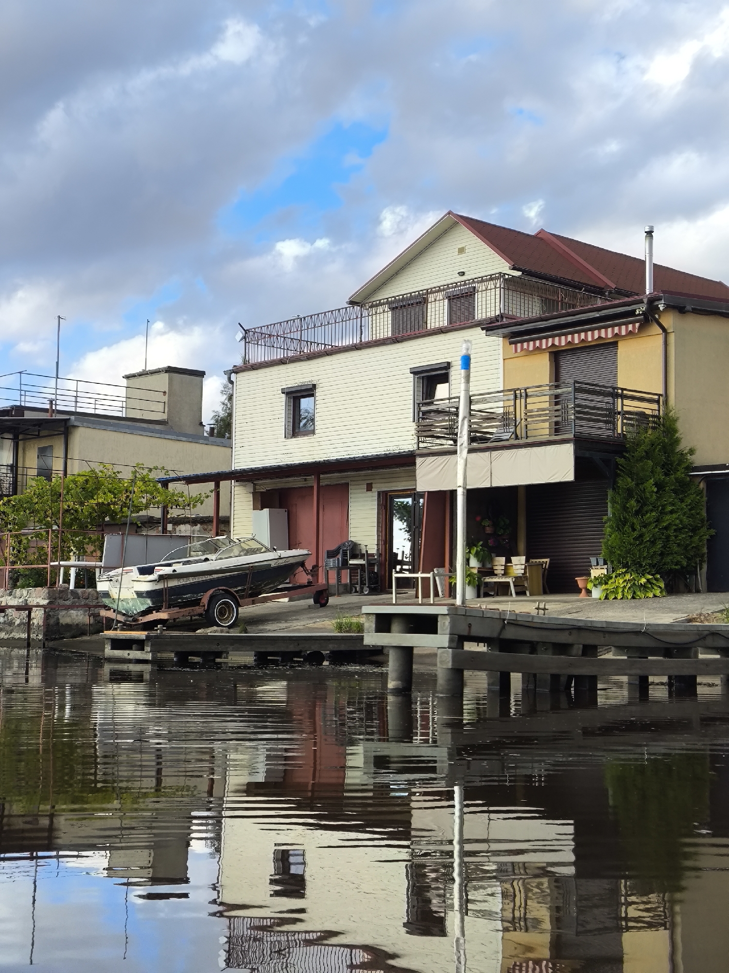 This is a forensic, matter-of-fact description of a scene located in Ushakovo, Russia. The image captures a waterside property with multiple structures and a boat.
In the foreground, a body of dark water reflects the scene above. A wooden dock, supported by vertical posts, extends into the water.
On the shore, a boat with a dark hull and white trim rests on a trailer. The boat appears to be a speed boat or runabout, with a visible windshield and seats. The trailer has two wheels and a rusted frame.
Behind the boat, a two-story building with a light-colored, horizontal siding is visible. This building has a dark-colored roof and a balcony with a metal railing on the upper level. Adjacent to this, a narrower, yellow-colored building has an awning with red and white stripes over its windows and a balcony with a black metal railing. A tall, slender evergreen tree stands next to the yellow building.
Further back, additional structures with flat roofs and windows are visible, suggesting a waterfront development. A chimney pipe protrudes from one of the buildings. The sky above is partially cloudy, with patches of blue sky visible through the gray clouds. The overall environment suggests a residential or recreational area situated near water. No people are visible in the image.