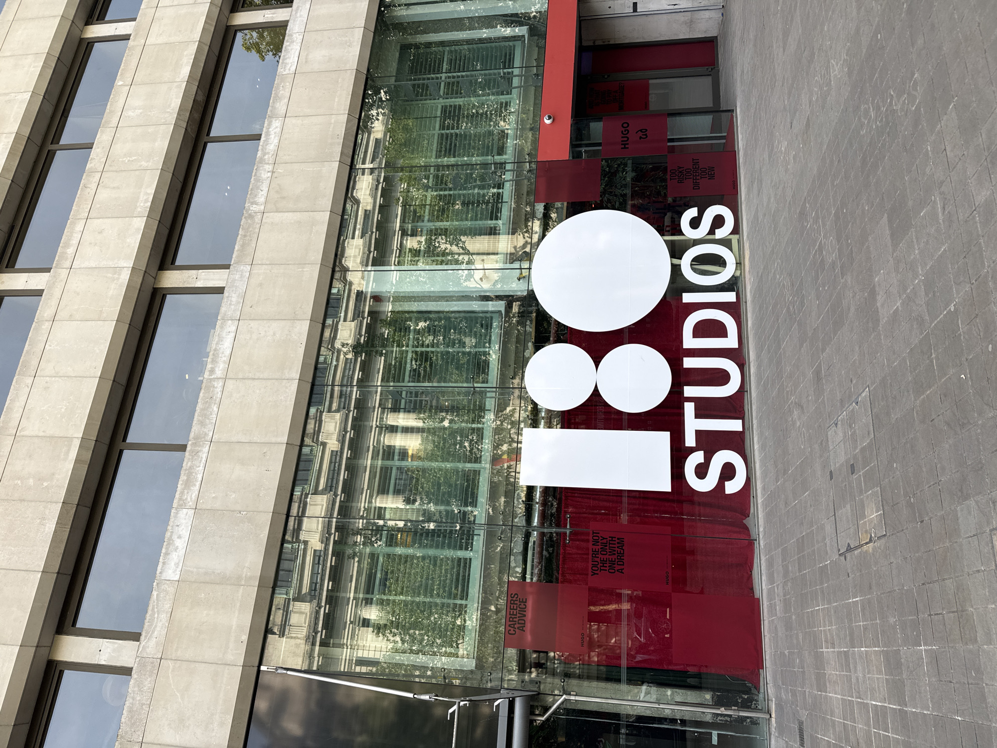 The image shows the storefront of "180 Studios" in London, United Kingdom. The building has a modern architectural style with large glass windows and stone cladding. The name "180 Studios" is prominently displayed in large white letters on the glass facade, with additional red signage indicating "HUGO" and related slogans such as "YOU'RE NOT THE ONLY ONE WITH A DREAM". The reflection in the glass shows a building across the street, suggesting an urban environment. The sidewalk is made of gray brick.
