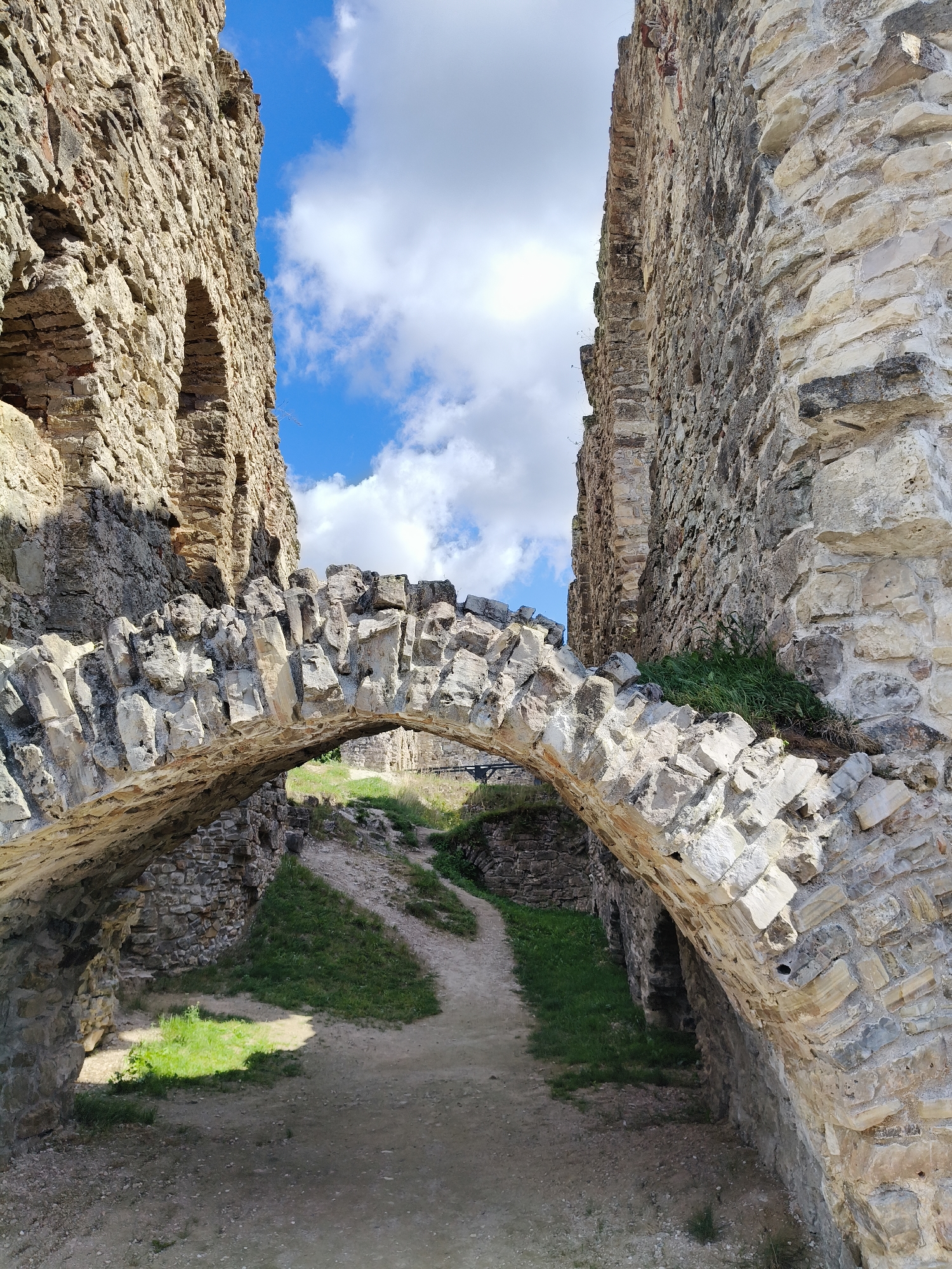 The image shows an arched stone bridge within the ruins of Koknese Castle in Koknese, Latvia. The bridge is constructed from rough, irregular stones and connects two crumbling stone walls of the castle. The walls are made of large, weathered stones, with visible texture and signs of age. Above the walls, a bright blue sky with scattered white clouds can be seen. A dirt path leads under the arch, with patches of green grass growing along its edges. The scene is bathed in sunlight, casting shadows that highlight the textures of the stone. The overall impression is one of historical ruins in a natural setting.