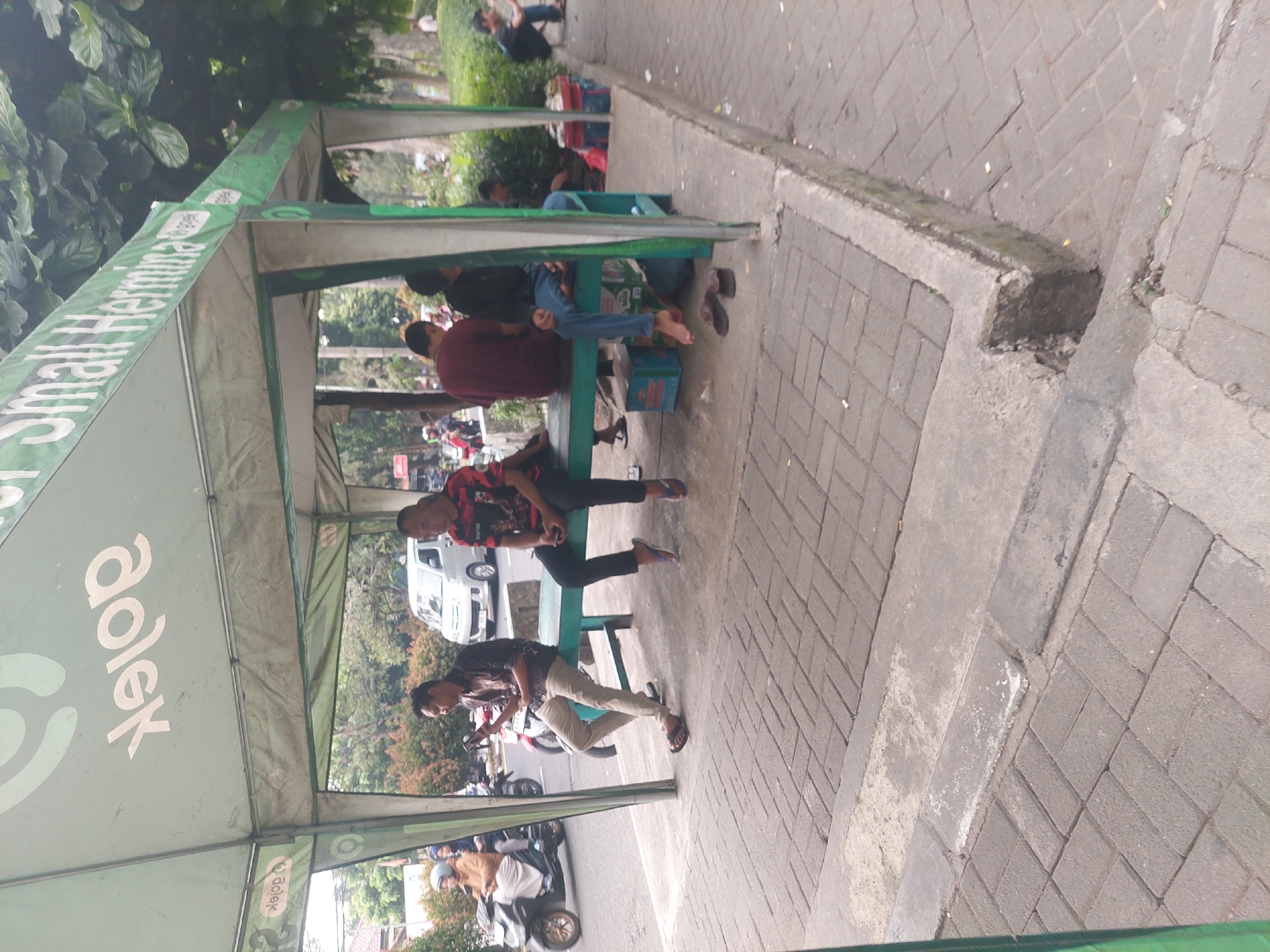 A group of men and women are gathered under a green awning that displays logos for "Gojek" and "Hermina Hospitals." The awning is situated on a sidewalk next to a street, suggesting it might be a waiting area for transportation services or a medical facility. The people are casually dressed and some are sitting on a green bench. A few are looking at their phones. In the background, motorcycles and a white van are visible on the street, indicating a busy urban environment. The sidewalk is paved with interlocking bricks. The setting appears to be an outdoor space in the city of Bogor, Indonesia, given the context of Gojek and Hermina Hospitals.