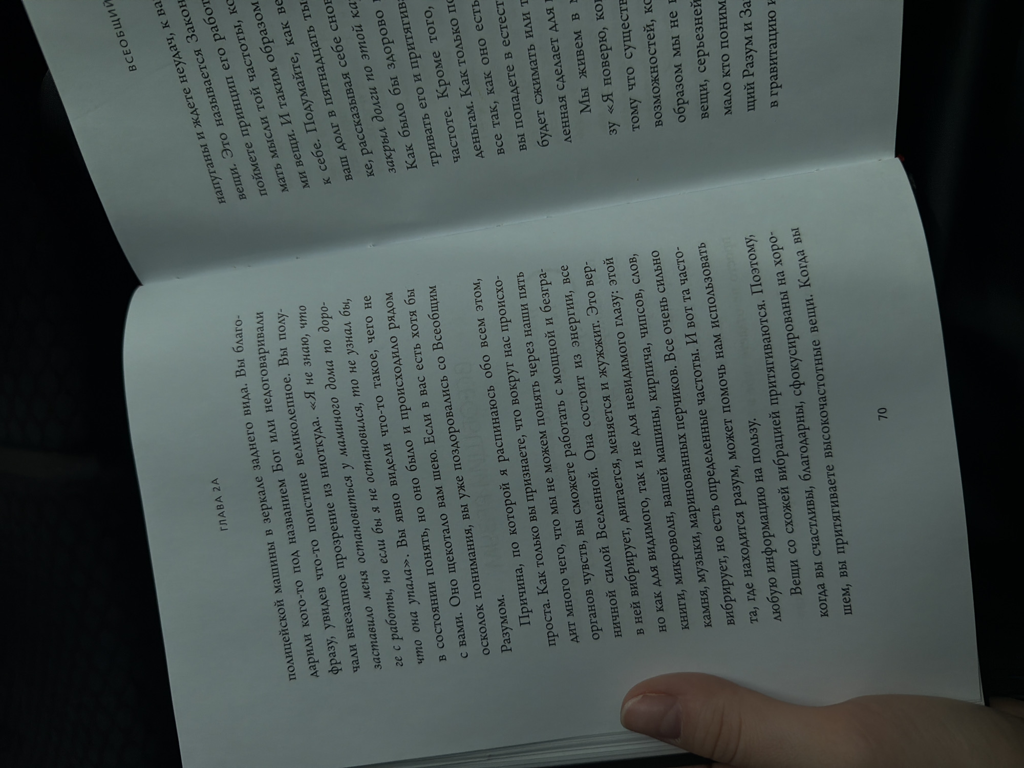 The image shows an open book with text in Russian. The book is lying on a dark surface, possibly a table. The text is printed in black ink on off-white paper. The left page has a chapter heading "ГЛАВА 2А" (Chapter 2A) followed by a paragraph of text. The right page has text that begins with "напуганы и ждете неудач, к ва" and continues downwards. The lighting suggests an indoor environment, possibly a library or a home. The image does not contain any elements that would indicate a specific location in Moscow, Russia.