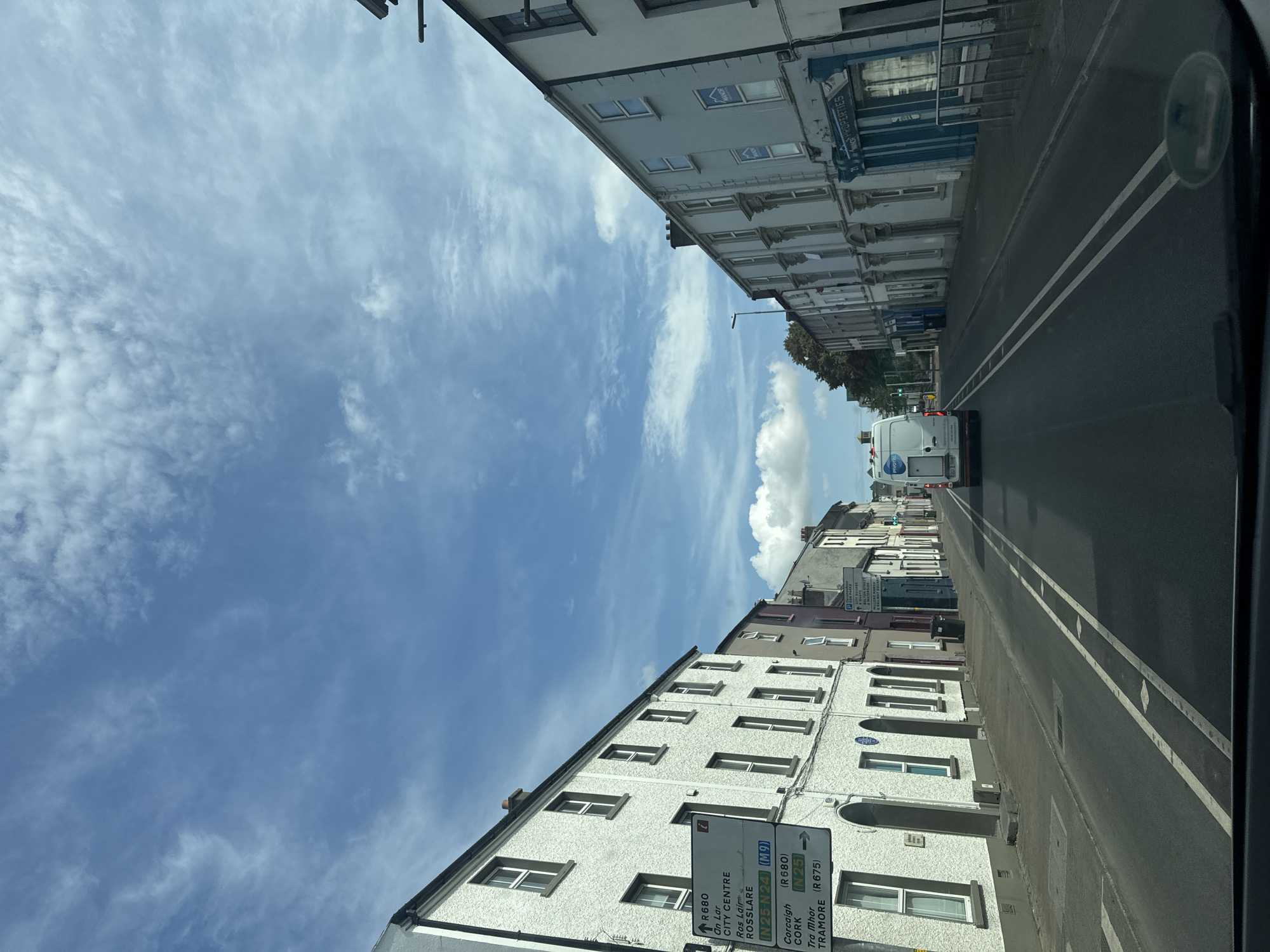 The image shows a street scene in Waterford, Ireland, looking down a road lined with terraced buildings. The buildings are predominantly white or pale-colored with sash windows. On the left side of the road, a white building with dark window frames is prominent, featuring a blue historical plaque and a white directional signpost. This signpost indicates directions to R680, On Lar, City Centre, Rosslare, N25-N24 (M9), Cork, and Tramore. Further down the street, on the right, is a row of buildings with shop fronts, one of which displays the name "MANOR PROPERTIES". A white van with "PIVOTAL" branding is driving away from the camera in the center of the road. The sky above is blue with scattered white clouds. The road has double white lines.