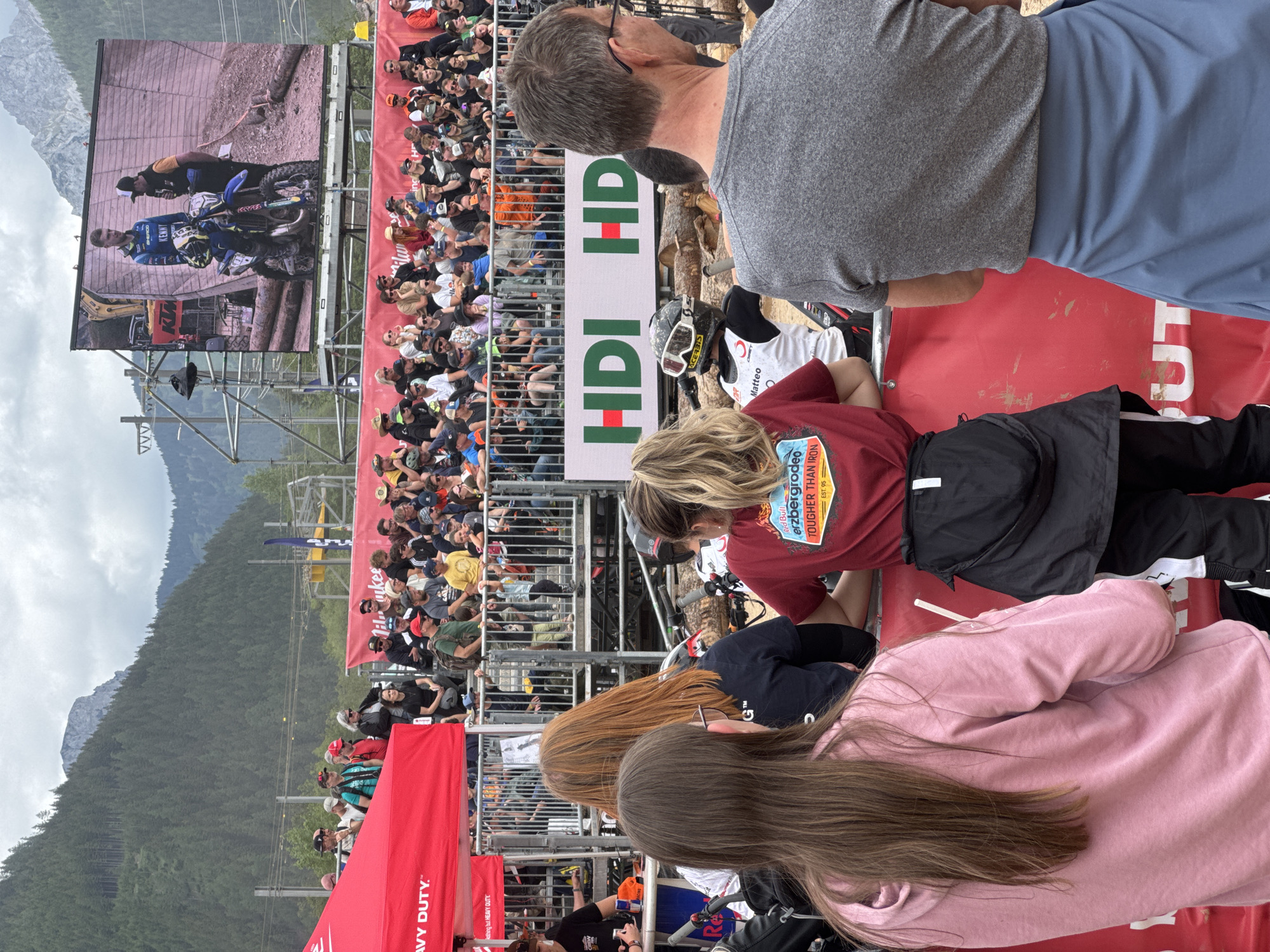This image depicts an outdoor extreme enduro event, specifically the Red Bull Erzbergrodeo in Eisenerz, Austria, under an overcast sky. In the background, steep, densely forested mountains rise, with a lighter, rocky peak visible in the distance on the upper right. The midground features a large spectator grandstand filled with numerous individuals of various ages, observing the event. Above the grandstand, a large LED screen displays a close-up of a male rider in blue motocross gear on a blue Sherco dirt bike, interacting with another man in a black jacket and cap. The rider's jersey prominently displays the name "KENNY." Utility lines and scaffolding supporting the screen are visible behind the grandstand. Below the grandstand, multiple red banners are visible, including one with "MILWAUKEE" and another with "HDI HDI" in green and red text. In the immediate foreground, a red barrier separates the audience from the event area, which is characterized by dirt and large wooden logs arranged as obstacles. Four individuals are prominent with their backs to the viewer: on the far left, a young woman with long brown hair wearing a pink hoodie; next to her, a woman with long reddish-brown hair wearing a dark shirt with a white graphic on the back; to her right, a woman with long blonde hair in a ponytail wearing a red t-shirt featuring the "Red Bull Erzbergrodeo TOUGHER THAN IRON EST. 95" logo, with a black jacket tied around her waist; and on the far right, a man with short gray hair and glasses, wearing a gray t-shirt and a blue jacket tied around his waist. Partially visible in the lower center, between the foreground spectators, is a person wearing a motocross helmet with goggles and a white jersey bearing "Matteo" and "Acerbis," positioned on a dirt bike, facing away from the camera. The scene captures a moment of active spectatorship at an extreme sport event.