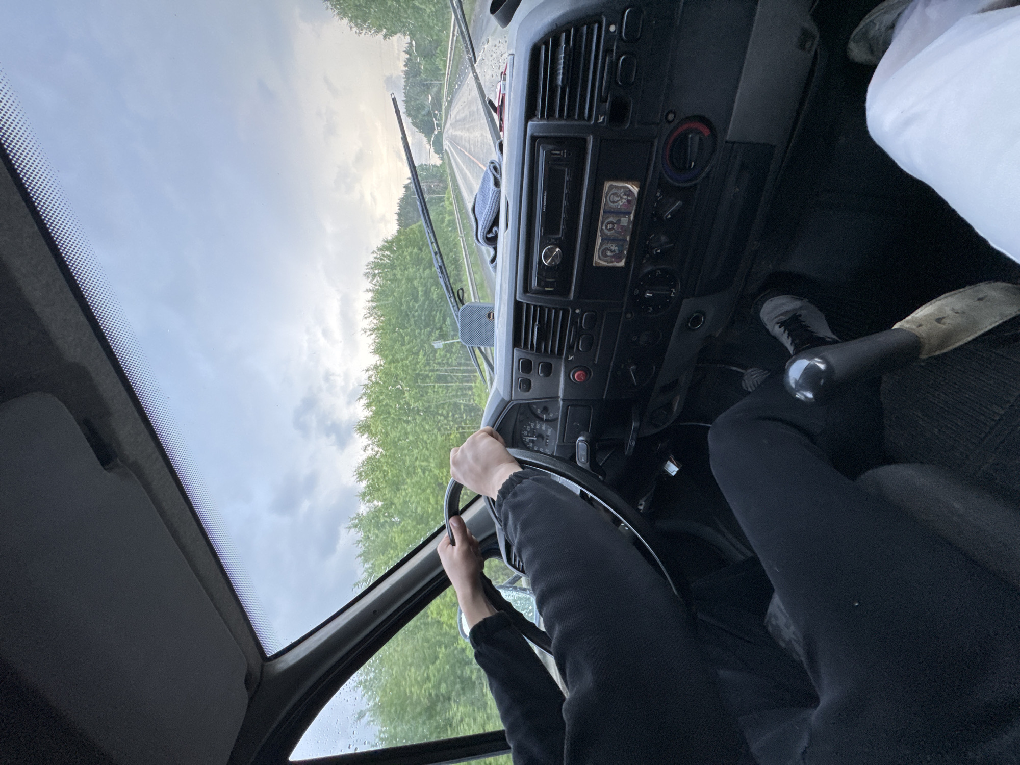 The image shows the interior of a van, viewed from the passenger's seat, looking forward. A person's hands are on the steering wheel, indicating they are driving. The dashboard is visible, with a radio, climate controls, and other indicators. The windshield shows a wet road and trees lining both sides, suggesting it is raining or has recently rained. The sky is overcast. The scene appears to be on a road outside of Pesterevo, Russia.