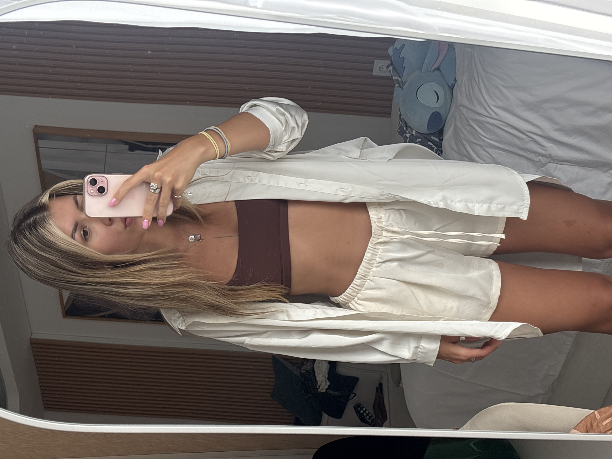 A woman is taking a mirror selfie in a room. She is wearing a brown bandeau top, white shorts, and an unbuttoned white oversized shirt. She has blonde hair and is wearing rings and bracelets. In the background, a bed with white sheets is visible, along with a blue Stitch plush toy. The room has wooden paneling on the wall. The lighting suggests it is daytime.