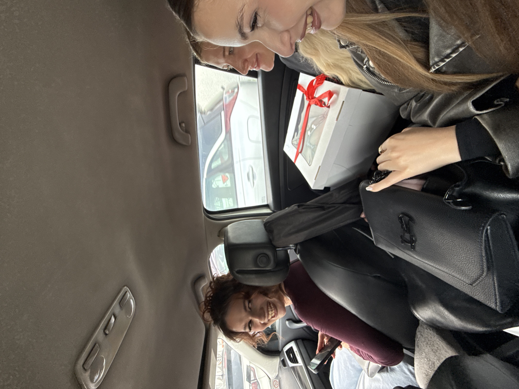 Two smiling women are inside a car. One is in the passenger seat with a white gift box adorned with a red ribbon and a black purse on her lap. The second woman is in the back seat, also smiling. Outside the car, other vehicles are visible. The location is likely within Vozdovac Urban Municipality, Serbia.