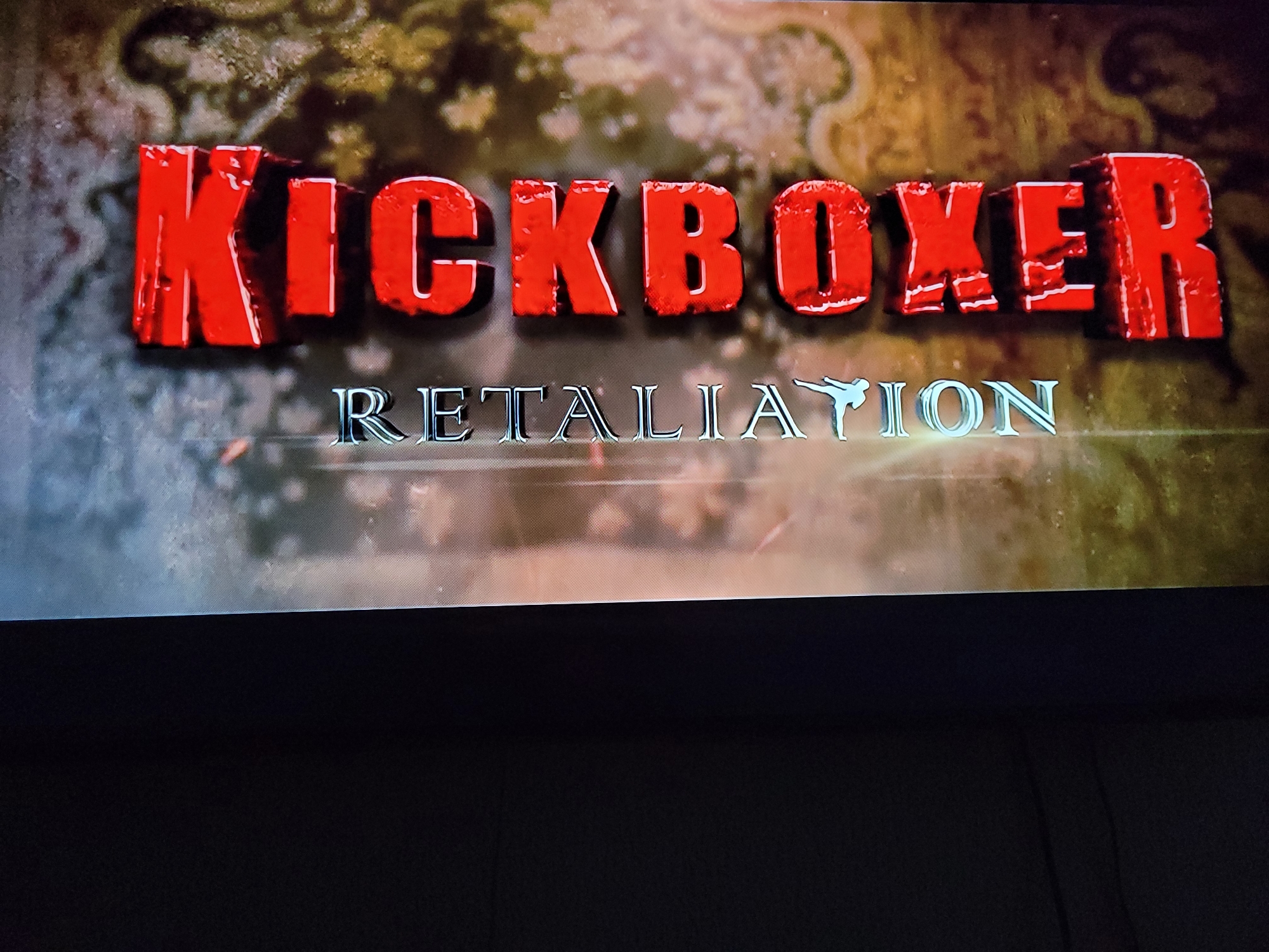 The image shows a movie title card for "KICKBOXER: RETALIATION". The title "KICKBOXER" is rendered in large, red, 3D block letters with a distressed texture. Below it, "RETALIATION" is written in a silver, metallic font, with a stylized silhouette of a kickboxer on the right end of the word. The background appears to be a textured, ornate pattern in muted brown and green tones. The scene is presented as if on a screen, with the bottom portion of the image being black, suggesting a cinema setting or a television screen. There are no people, objects, actions, or interactions visible in the scene, nor is there any identifiable location within a city or country.