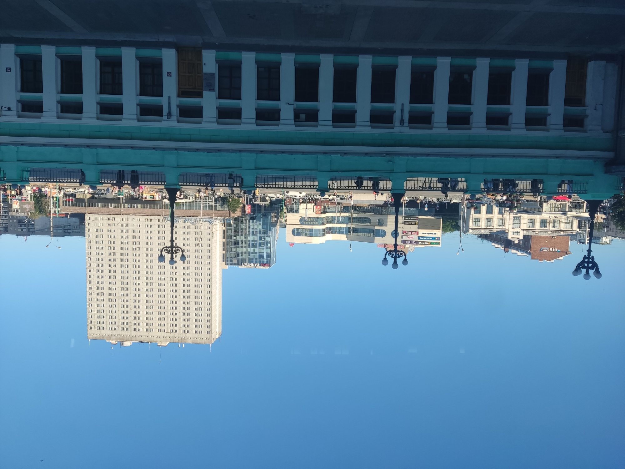 This is an upside-down photograph of a cityscape, likely taken during the day with clear, blue skies. The scene is dominated by several buildings of varying architectural styles. In the foreground, a large, light-colored rectangular building with numerous windows stretches across the frame. To its left, a cluster of modern glass-fronted buildings can be seen, some appearing to be office blocks or shopping centers. Further back, a lower, older building with a light green facade and white columns runs horizontally across the top of the image. Reflections of people and various objects, including cars and streetlights, are visible in what appears to be a polished surface below the buildings, suggesting the photograph was taken from a reflective surface, possibly a window.
Several signs with text are partially visible. One sign reads "CAPITAL" in large letters. Another appears to say "rgent" and "zella". A red sign shows Cyrillic text, with "БТБ БАНК" being the most legible part, which translates to "BTB BANK". There are also some smaller signs with what looks like brand names or shop names. The overall impression is that of an urban environment, possibly a commercial or business district. The clear sky and bright lighting suggest it is a pleasant day, though the upside-down perspective creates a disorienting visual effect.