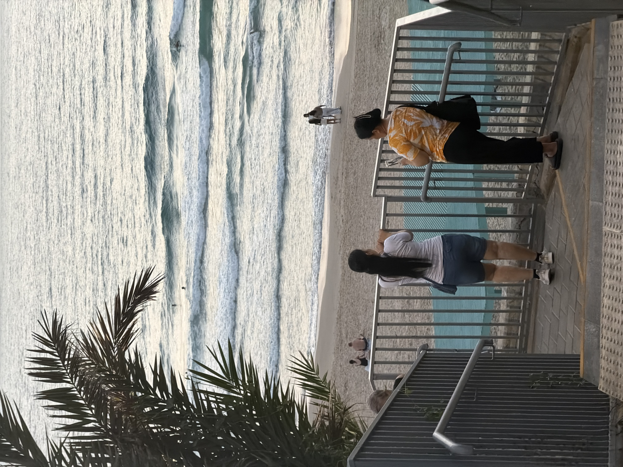 The image depicts a view from a pathway overlooking a beach in Bat Yam, Israel. In the foreground, two women are standing near a metal railing. The woman on the left has long dark hair and is wearing a white t-shirt and denim shorts. The woman on the right has dark hair tied up and is wearing an orange and white patterned shirt and black pants. Both are looking out towards the sea. In the middle ground, several people are visible on the sandy beach. Some are sitting, and others are standing and walking. The sea has moderate waves with whitecaps, and a few individuals can be seen in the water, possibly swimming or surfing. A palm frond from a nearby tree extends into the upper left corner of the frame, casting a shadow over a portion of the scene. The lighting suggests it is either early morning or late afternoon, given the soft light on the water.