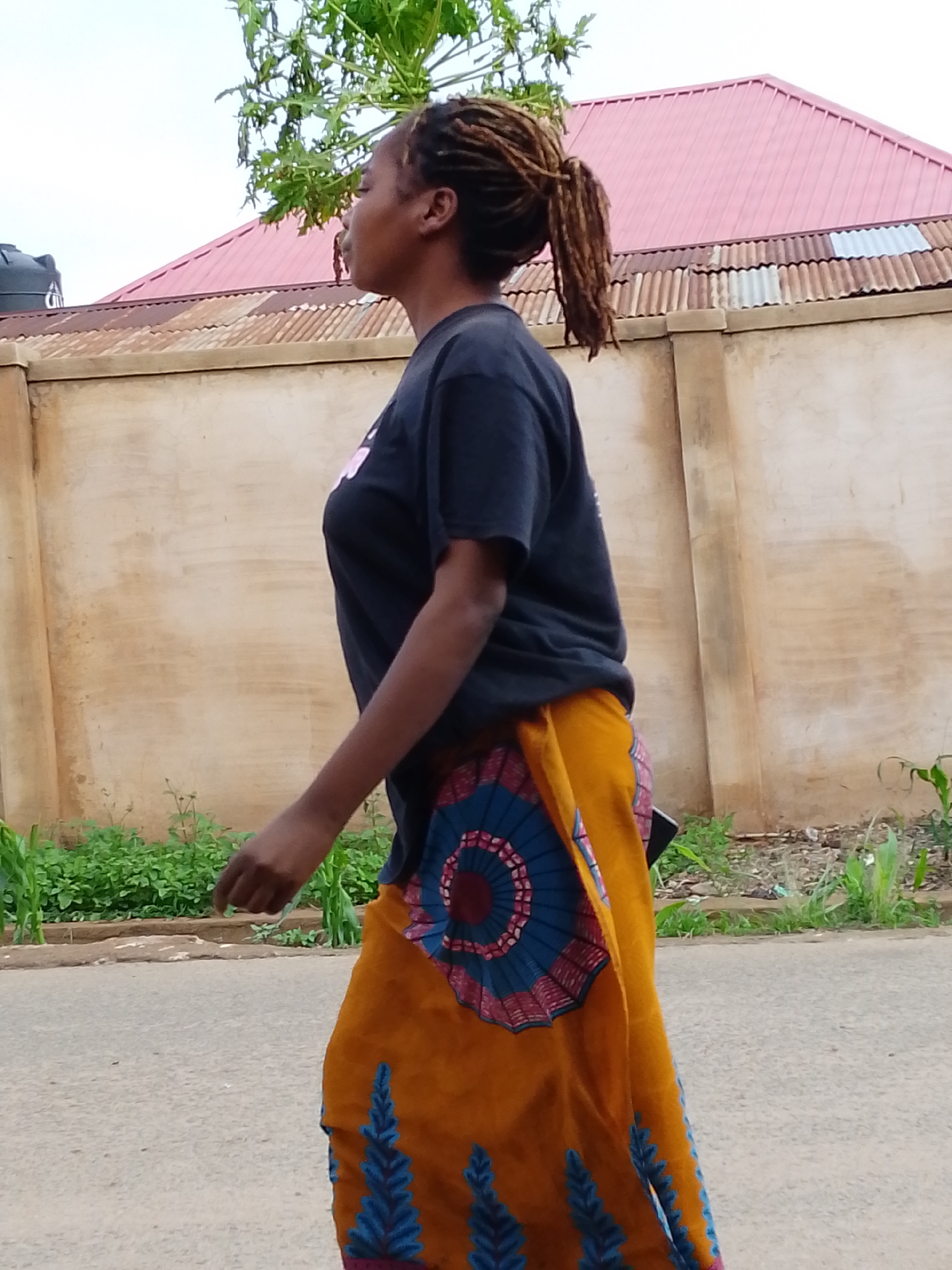 A woman is walking along a street in Jos, Nigeria. She is wearing a black t-shirt and a long, yellow skirt with blue and red patterns. Her hair is styled in dreadlocks pulled back into a ponytail. The background shows a wall with a building behind it featuring a pink roof. There is greenery along the base of the wall, and a street is visible in the foreground.
