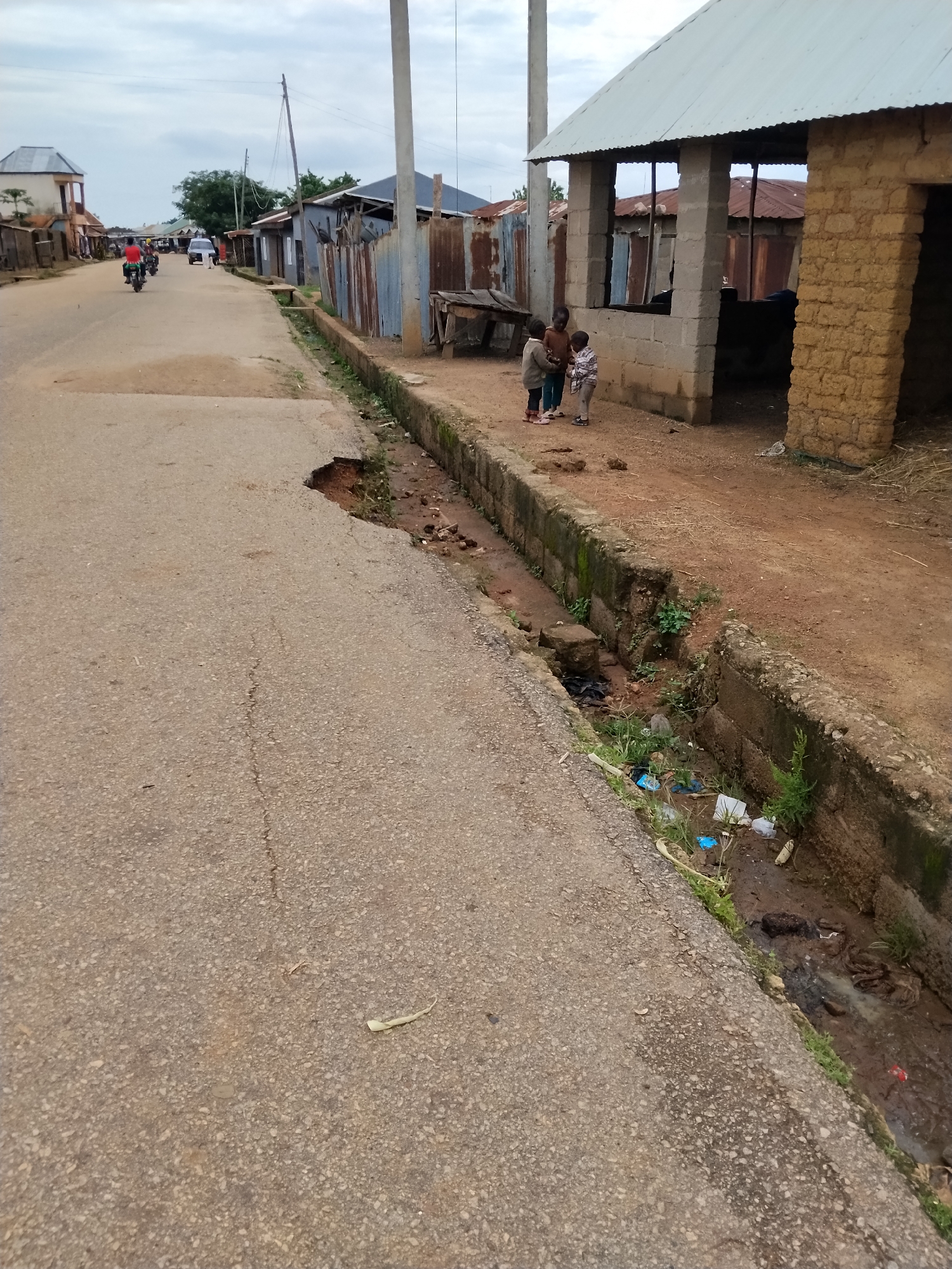 The image depicts a street scene in Mangu Town, Nigeria. A paved road, showing signs of wear and a significant pothole on the left side, runs diagonally through the frame. Adjacent to the road is a concrete drainage channel, partially filled with debris, including plastic bottles and other refuse. On the right side of the drainage channel, there is a building constructed with mud bricks and a corrugated metal roof. Three young children are standing near this building, looking towards something out of frame. Further down the road, there are other structures, some with corrugated metal roofs and tin fencing. In the distance, several motorcycles and a car are visible on the road, suggesting ongoing activity. The sky is overcast.