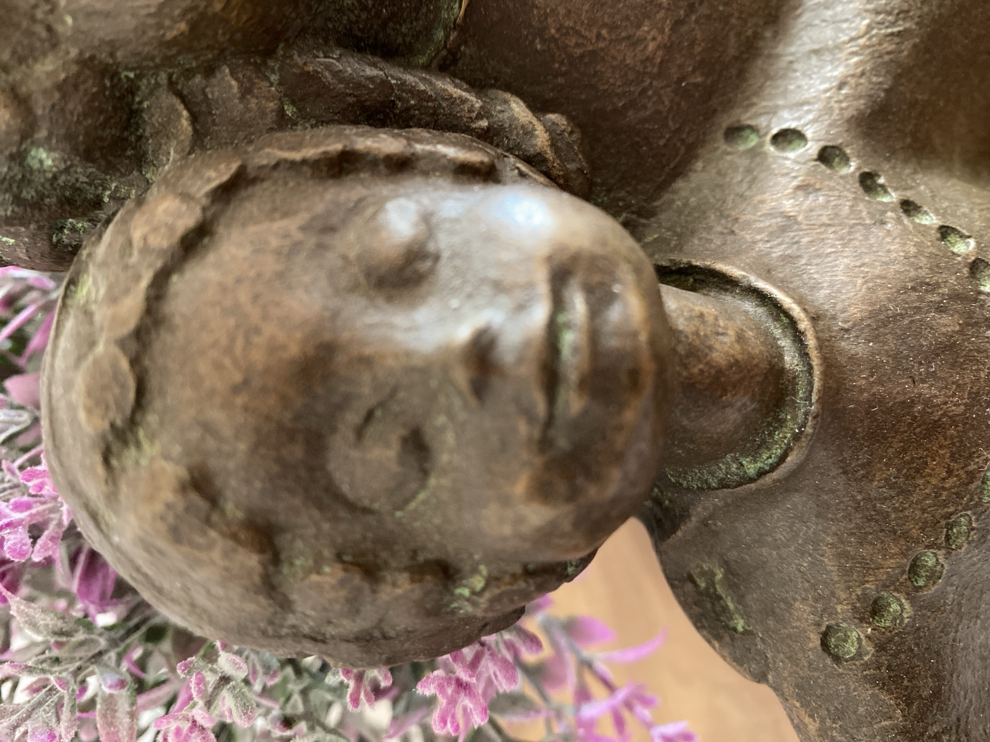 A close-up shot captures a bronze statue's head resting on purple flowers. The statue appears aged with patina and distinct facial features, suggesting a classical style. No specific location in Budva, Montenegro is discernible.