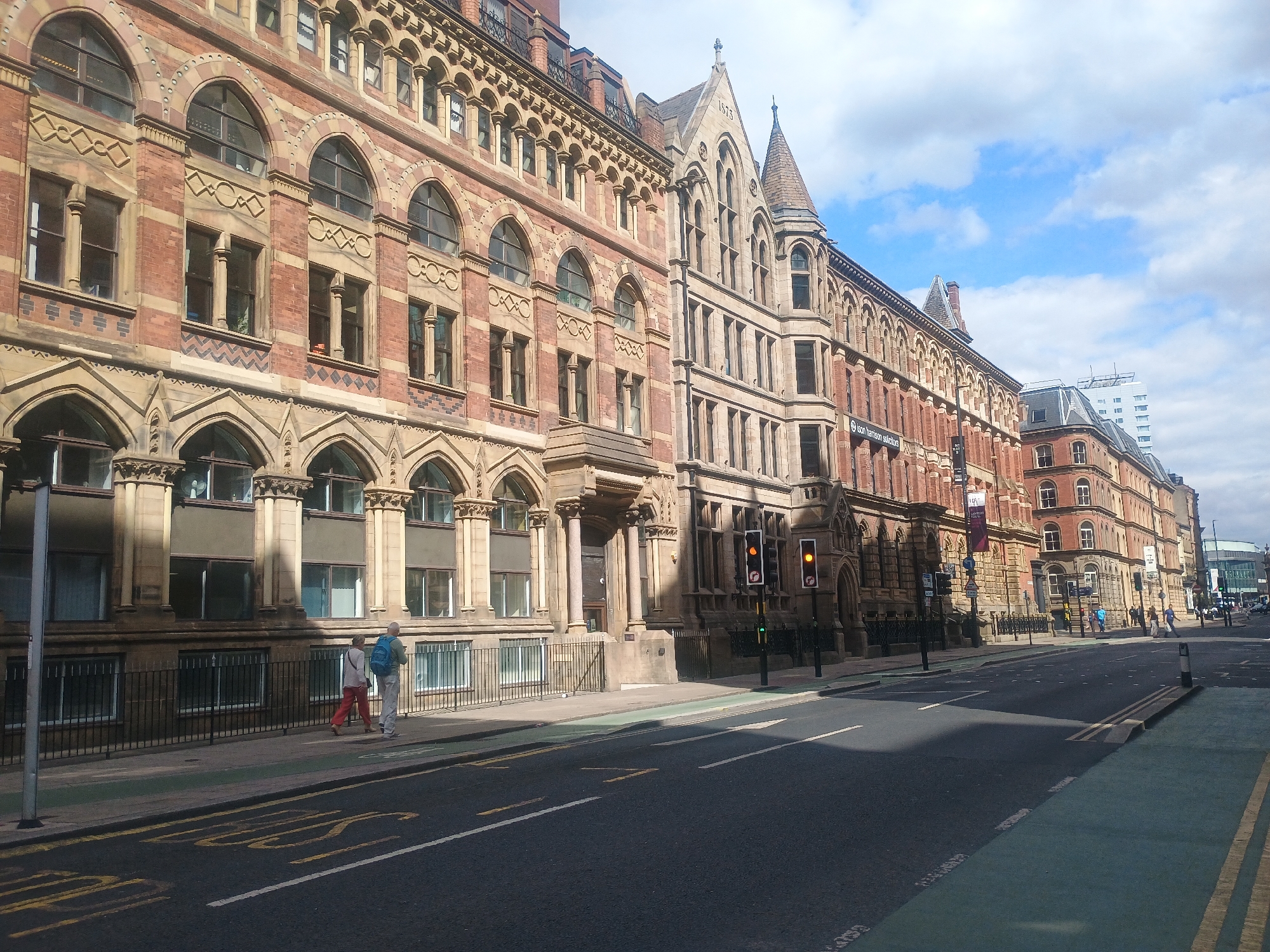 The image shows a street scene in an urban environment with large, ornate brick buildings lining the street. The architecture of the buildings features arched windows, decorative stonework, and pitched roofs. There are several pedestrians walking on the sidewalk. The street is paved and has lane markings, including a bus lane indicated by green paint. Traffic lights are visible, showing orange and red signals. A few cars are present further down the street. The sky is partly cloudy. The scene appears to be from a daytime setting.