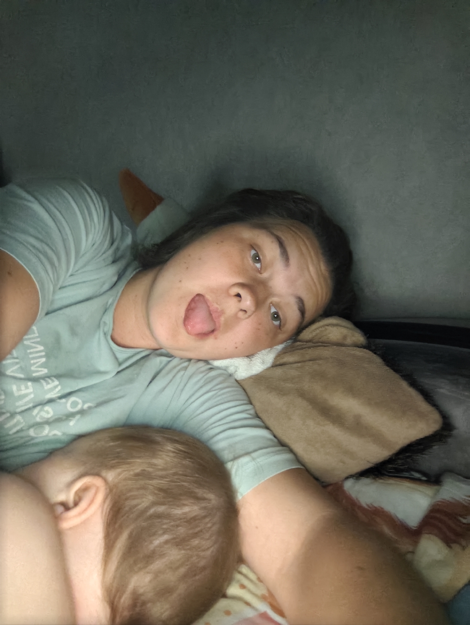 A woman lies on a bed next to a sleeping infant. The woman has dark hair and freckles and is sticking her tongue out. She is wearing a light green t-shirt. The infant has fair hair and is facing away from the camera. The bed has a patterned blanket and a brown pillow. The location is in an indoor room with grey walls. The photo has an amateur quality, and the city of Chaikovsky, Russia cannot be determined.