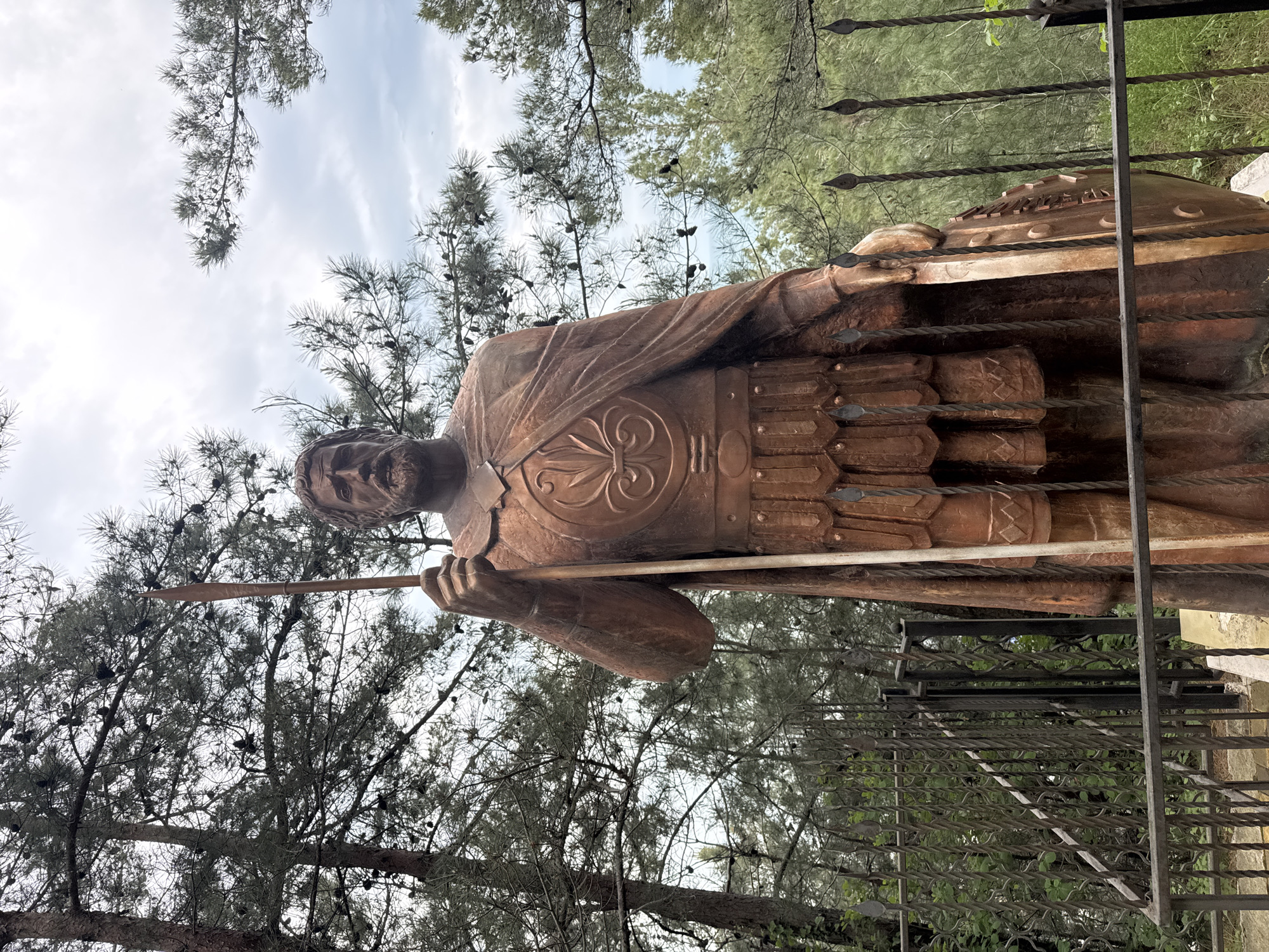 The image shows a bronze statue of a warrior holding a spear, possibly in Sosnovy Bor, Russia. The statue is partially obscured by tree branches and stands behind a decorative metal fence. The sky is overcast with white clouds. The warrior is wearing a breastplate with a stylized floral design and a kilt-like garment.