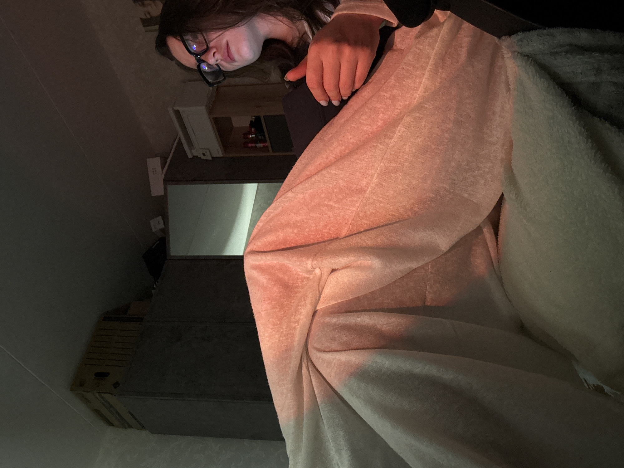 A young woman wearing glasses is partially visible in the upper-right corner of the image. She is covered with blankets that appear to be two different colors - a pinkish-white blanket on the left, and a fluffy gray blanket on the right. The room contains shelves loaded with boxes or items in the background. Electrical outlets are affixed to the wall, and there appear to be pipes or other fittings going up the walls. The location and context of the image suggest that it may have been taken in a residential space, but more information is needed to confirm the specific location in Chaikovsky, Russia.