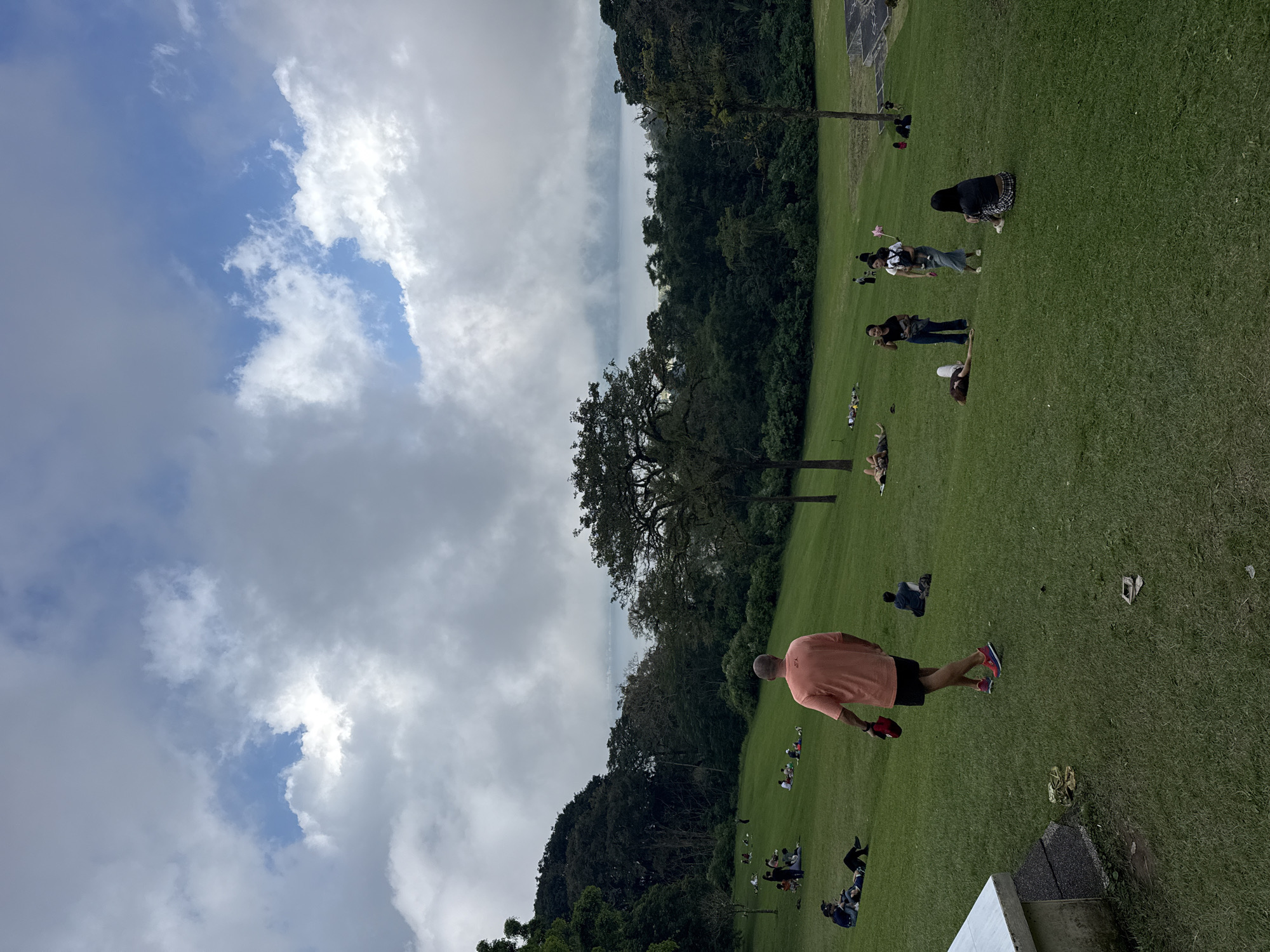 The image depicts a grassy field in Baturiti, Indonesia, under a partly cloudy sky. A line of trees and mountains forms the horizon. Several people are scattered across the field, some lying down, others sitting or standing. A man in a peach-colored shirt and black shorts is walking across the field.