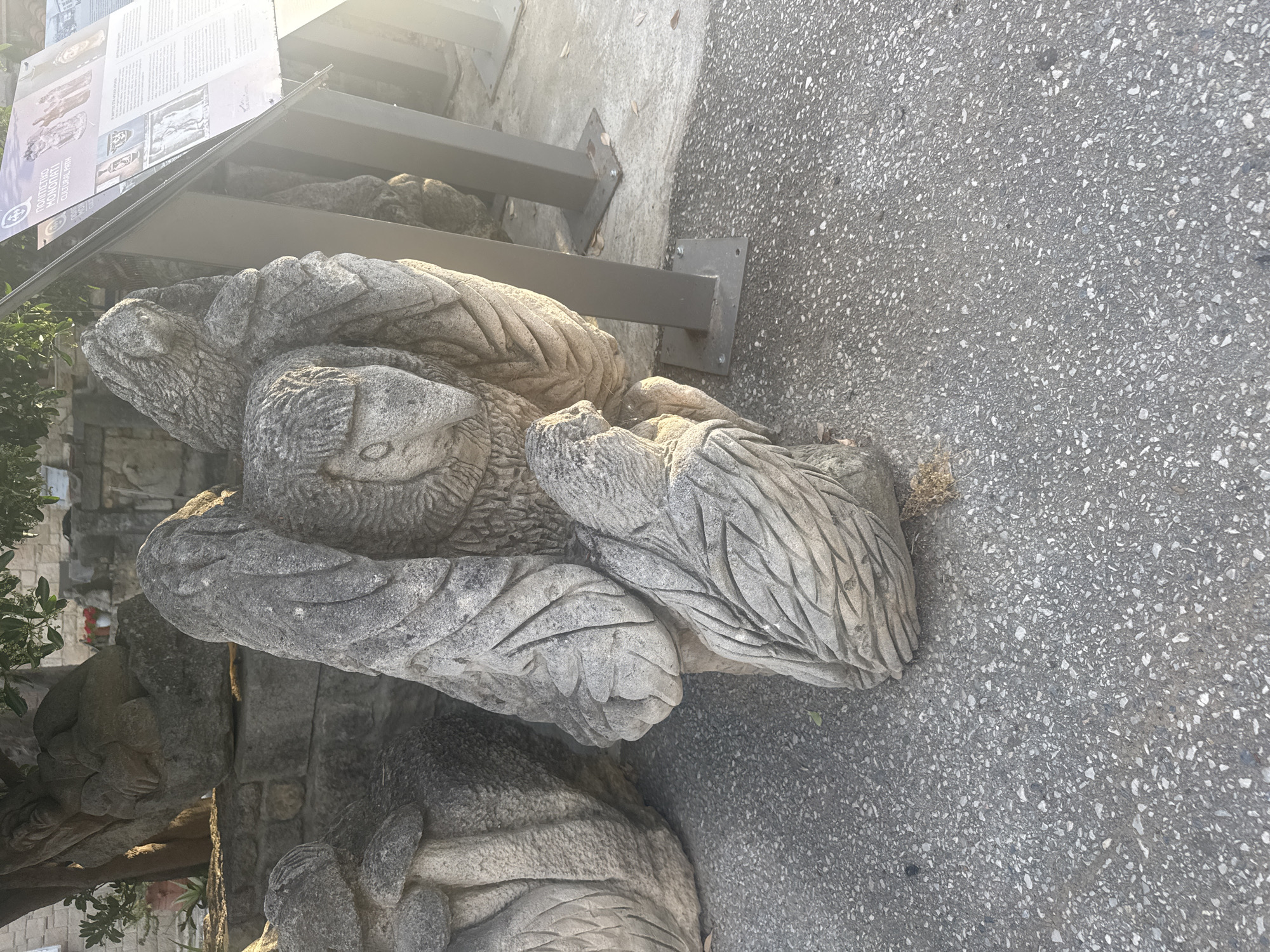 An eye-level, outdoor shot captures a stone sculpture of what appears to be a bird or owl with its wings spread and a smaller creature nestled underneath. The sculpture is made of rough-hewn stone, with visible chisel marks creating texture and detail on the feathers and faces. Behind the main sculpture, other stone elements and a tree are visible, suggesting an artistic installation or park setting. To the right of the sculpture, a metal information stand with text and images is partially visible. The ground is paved with asphalt. This artwork is located in the Municipal Unit of Kassandra, Greece.