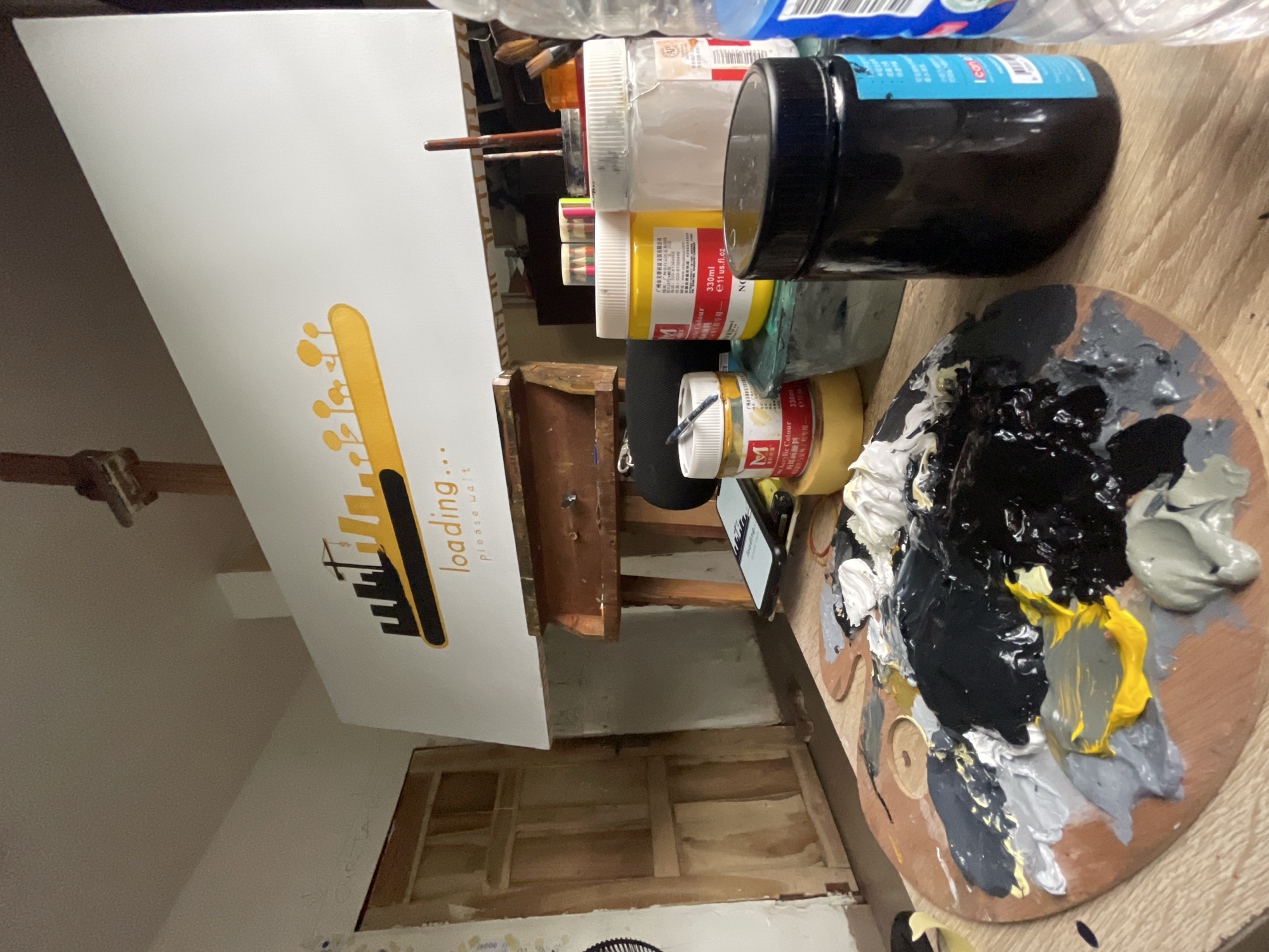 The image shows an art studio setup. A partially completed artwork is displayed on an easel, featuring a graphic design with the word "loading..." written on it. Next to the easel is an artist's palette with several colors of mixed paint, including black, white, grey, and yellow. Paint containers are stacked nearby. The floor and walls appear unfinished, exposing wooden structures. The location is not discernible from the image.