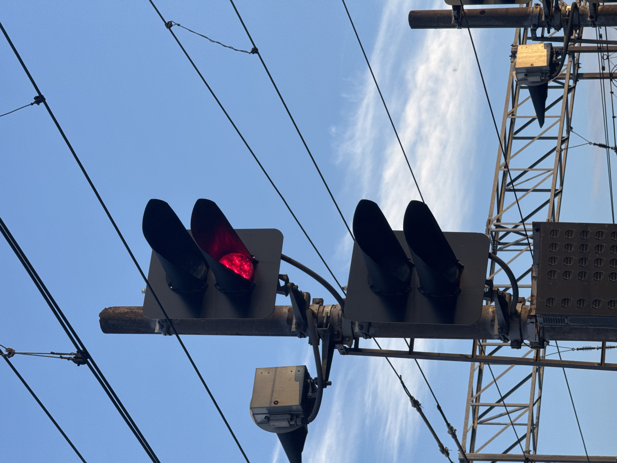 This image shows a railway signal displaying a red light, situated in Saint Petersburg, Russia. The signal pole is a dark, vertical metal structure, with multiple horizontal wires suspended above and around it, likely part of the railway's overhead electrical system. A boxy, grey electrical component is attached to the left side of the pole. Below the top signal, there is another signal housing containing two dark, unlit signal lights. In the background, a complex metal structure, characteristic of railway infrastructure, is visible. The sky is a clear blue with wispy white clouds.