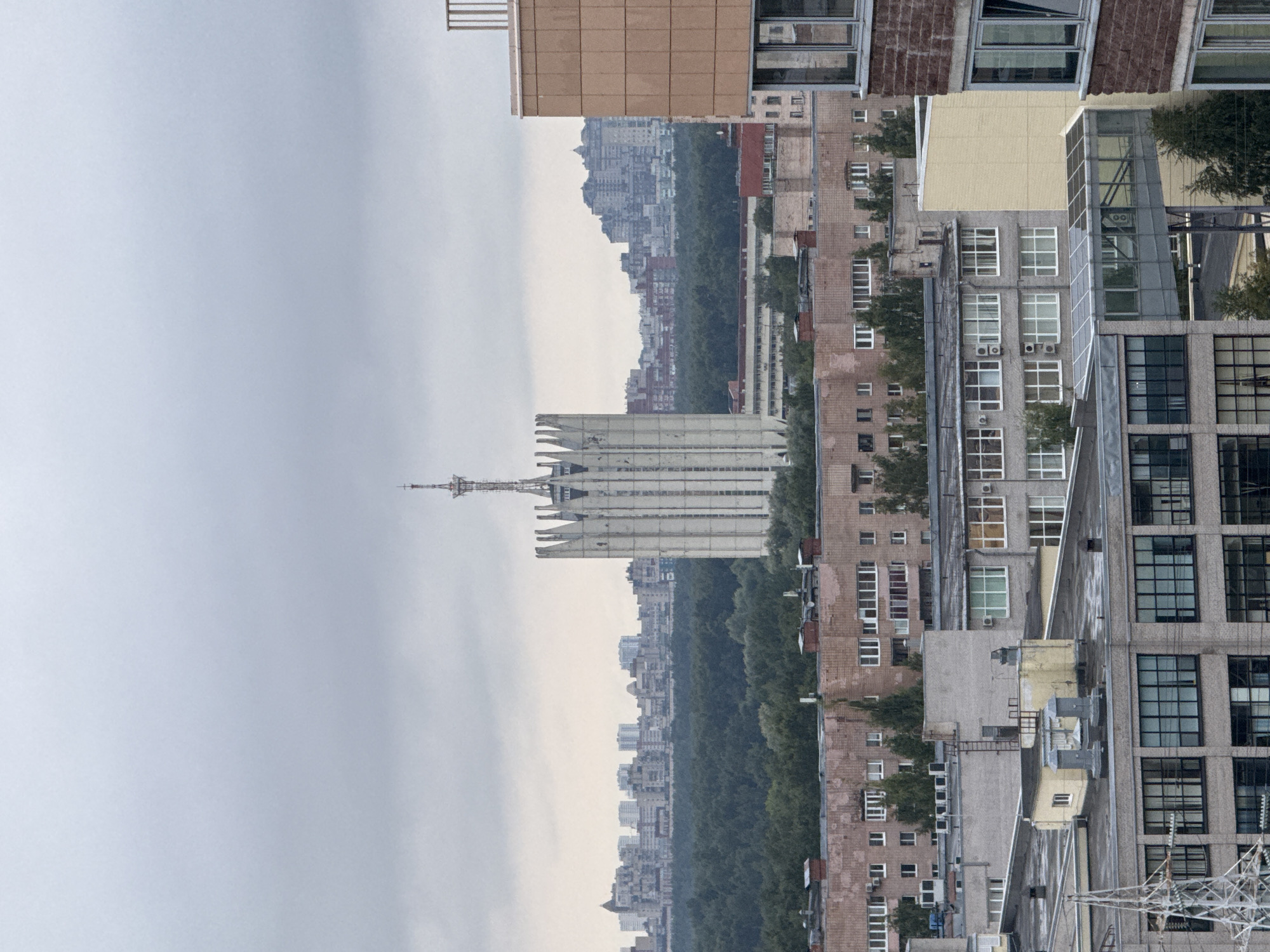 The image shows a panoramic view of a cityscape under an overcast sky. In the foreground, several multi-story residential buildings with brick and concrete facades are visible, some with visible air conditioning units and satellite dishes on their roofs. A large, modern-looking glass-enclosed walkway connects two of these buildings. Behind these buildings, a dense forest stretches across the mid-ground.
Dominating the middle ground is a distinctive, tall, monolithic concrete building with a pointed, crenellated top and a radio tower on its summit. This structure appears to be a prominent landmark. Further in the background, a sprawling urban area with numerous apartment buildings of varying heights and architectural styles is visible, some of which are set against a hazy, distant skyline. The overall impression is of a moderately developed urban area with a mix of residential, industrial, and possibly administrative buildings, set against a backdrop of greenery and a cloudy sky. The image appears to be taken from an elevated vantage point, looking out over the city.