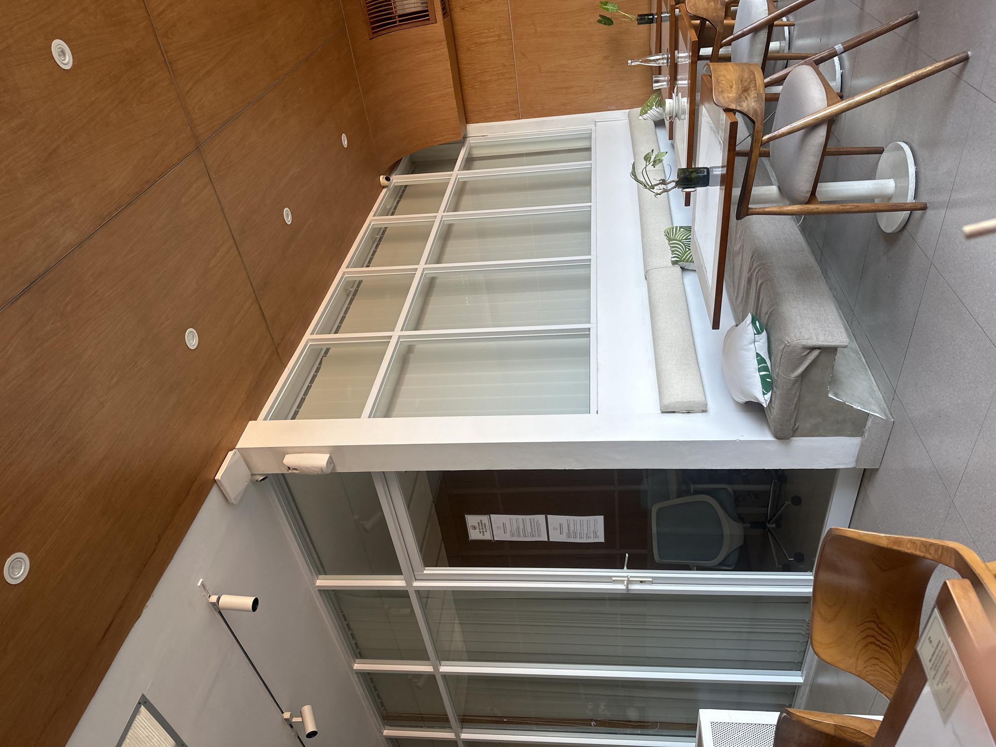 The image shows the interior of a modern cafe in North Kuta, Indonesia. The cafe features a wooden-paneled ceiling with recessed lighting and a white, modern aesthetic for the walls and furniture. The seating area is divided by a glass partition, revealing a separate room with a desk and office chair. The main cafe area has tables with wooden chairs and a long, built-in bench with cushions and pillows. Plants are used as decorative elements on the tables. The overall impression is one of a clean, minimalist, and contemporary dining space.