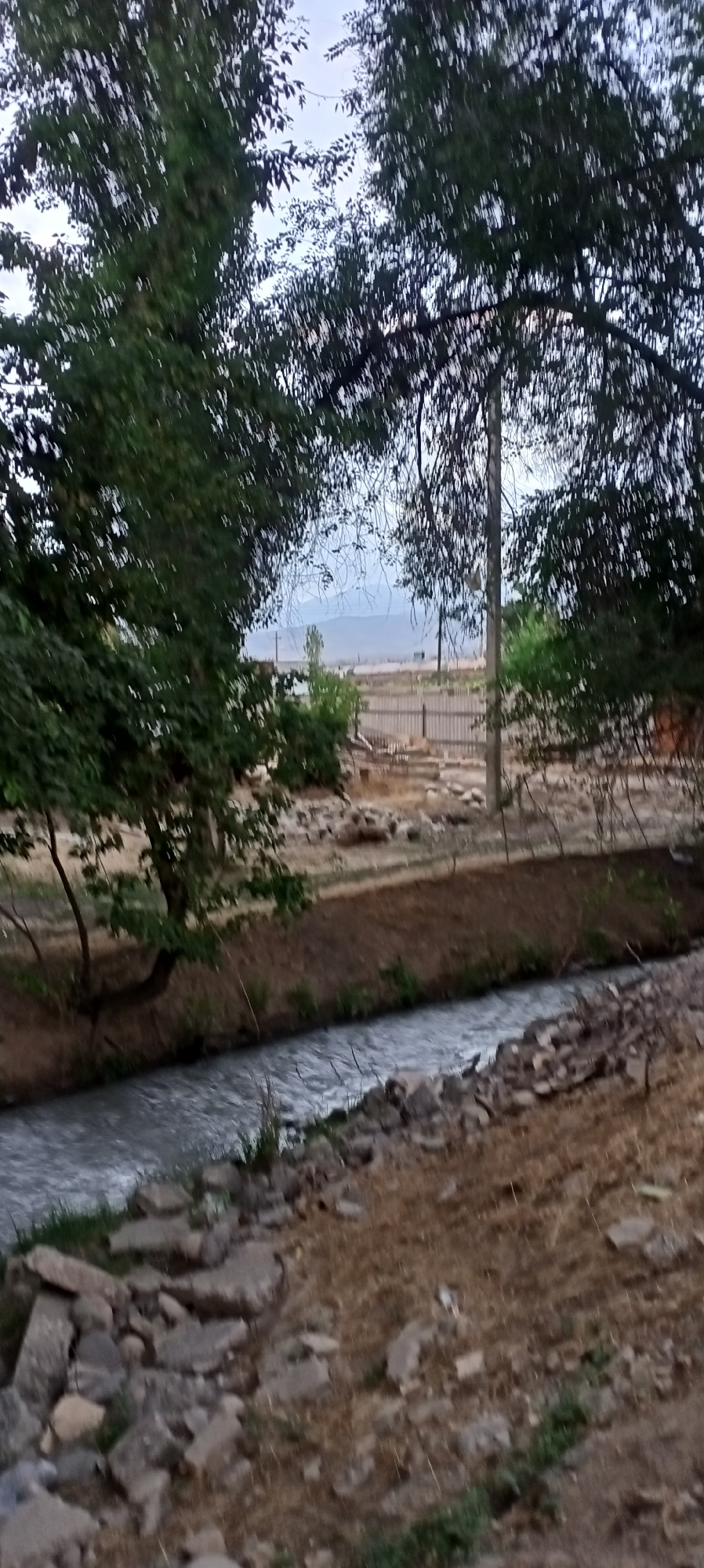 The image depicts a tranquil, natural scene in Sokuluk, Kyrgyzstan. In the foreground, a small, shallow stream flows, its surface reflecting the overcast sky. The banks of the stream are uneven, with scattered rocks and dry vegetation. To the left of the stream, a large tree with dense green foliage casts a shadow, its trunk visible and leaning slightly. Beyond the stream, a dry, dusty area is populated with more rocks and sparse, dry grasses. Further in the background, a wooden fence runs horizontally, with what appears to be a few sparsely vegetated structures or buildings visible behind it. A utility pole stands near the fence. In the distance, hills or mountains are faintly visible under a hazy sky. The overall impression is one of a rural or semi-rural setting on a cloudy day. There are no people or animals visible in the image.
