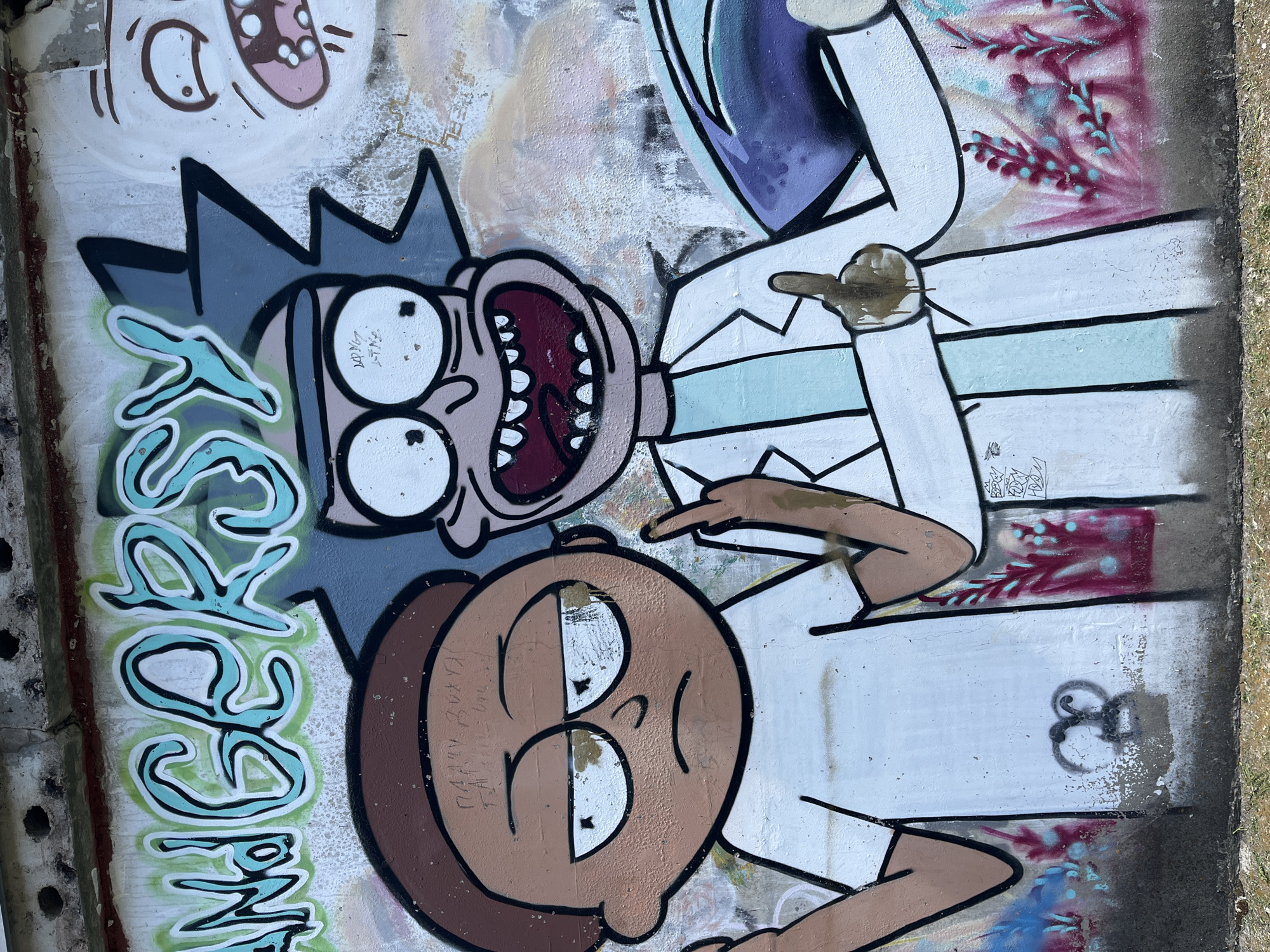 The image is a photograph of graffiti on a concrete wall. The graffiti depicts cartoon characters resembling Rick Sanchez and Morty Smith from the animated television series "Rick and Morty." Rick, on the right, is wearing a lab coat and has a wide, manic grin. Morty, on the left, has a grumpy expression and is wearing a white t-shirt. Both characters are pointing fingers. Above them, in large, stylized lettering, are the words "ANGERSY." The graffiti is colorful, with teal, green, white, and black as the dominant colors. The wall itself appears weathered and has some peeling paint and dirt. The background shows a rough concrete texture and some patchy green and red spray paint that suggests foliage or abstract patterns. The ground in the foreground is a mix of dirt and grass. This graffiti is located on the exterior wall of the Votkinsk Hydroelectric Power Plant in Chaikovsky, Russia.