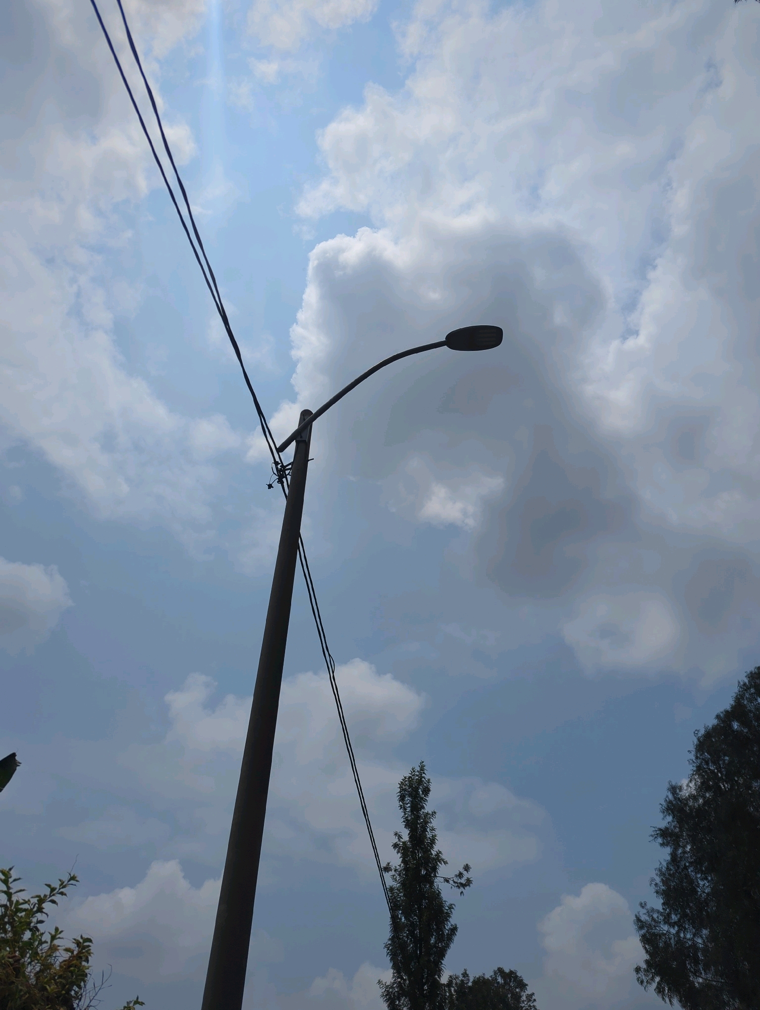 A low-angle, full shot captures a light pole silhouetted against a bright, partly cloudy sky. The light pole, made of dark material, stands vertically off-center to the left, its top extending out of frame. A street lamp is attached to the pole, facing right. Two dark wires run diagonally across the upper portion of the frame, connecting to the pole. In the lower right corner, the dark, feathery branches of a tree are visible against the sky. The sky is a pale blue, with large, fluffy white clouds dominating the upper and right portions of the image. The sun is partially obscured by a cloud, creating a bright, diffused light source in the upper left quadrant of the frame. The scene appears to be outdoors, possibly in an urban or suburban setting within Musanze District, Rwanda, given the presence of a street light and overhead wires, common infrastructure in populated areas.