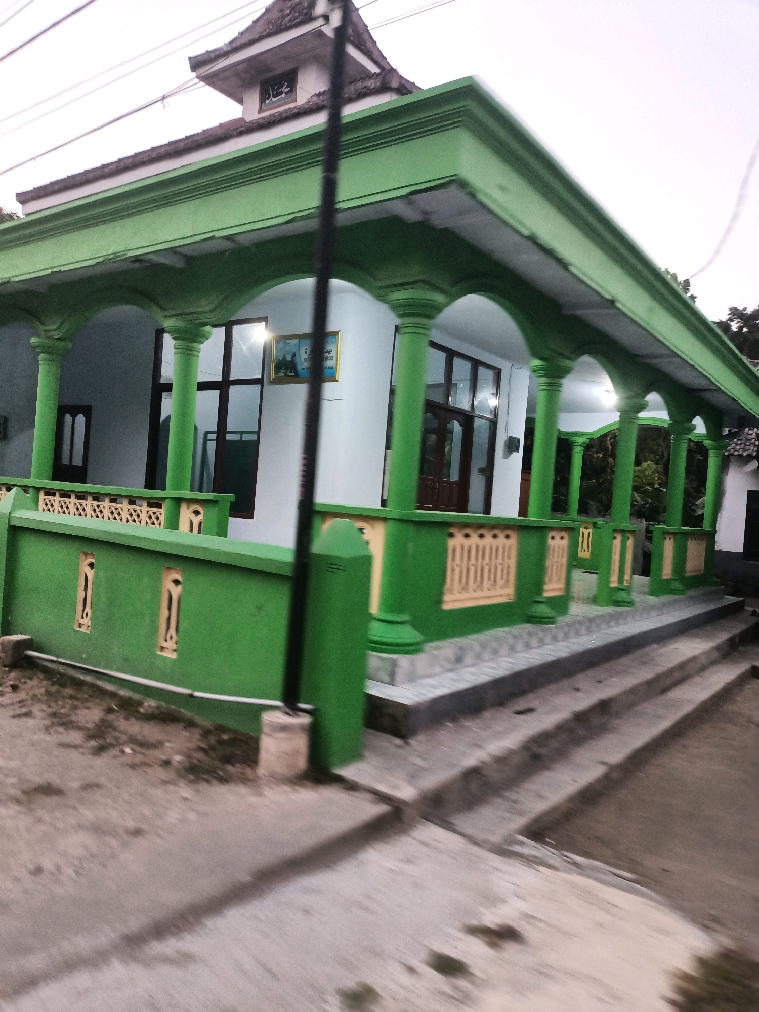 The image shows a green building with a traditional Indonesian architectural style, likely a mosque or a community center, located in Badas, Indonesia. The building features a prominent green roof with an extended overhang supported by numerous green pillars. A green, ornate fence with cream-colored detailing runs along the front of the property, separating it from a paved walkway and a dirt path. The walkway leads up a set of concrete stairs to the entrance of the building. The building's walls are white, and it has large windows with dark frames and wooden doors. A framed picture is visible inside one of the windows. The sky appears overcast. The image is taken from a slight angle, giving a sense of depth and perspective.