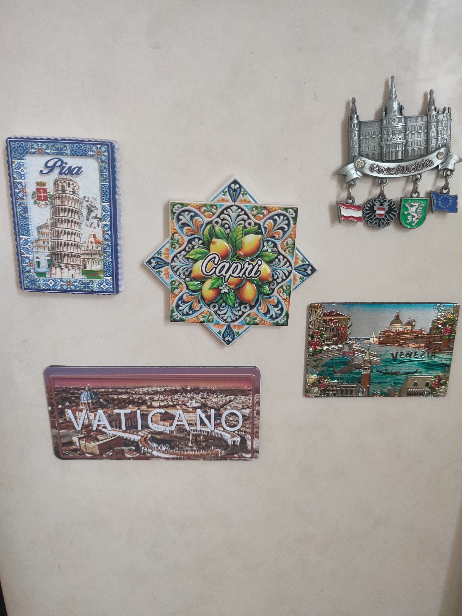 This is a close-up, eye-level, horizontal, indoor shot of a refrigerator door covered in magnets, taken in Kriukivshchyna, Ukraine. The background is a plain, light-colored refrigerator surface, showing faint smudges and dust.
Dominating the left side of the frame is a rectangular magnet featuring the Leaning Tower of Pisa, with the word "Pisa" prominently displayed at the top. Below it, an ornate, diamond-shaped magnet depicts lemons and colorful patterns, with the word "Capri" in the center.
To the right of the Capri magnet, a silver-colored metal magnet represents a building with intricate architectural details and a banner reading "Graz Austria". Dangling from this main magnet are four smaller shield-shaped magnets, each displaying a different country's flag or crest.
Below the Pisa and Capri magnets, a rectangular magnet shows an aerial view of Vatican City, Rome, with the word "VATICANO" emblazoned across it in large, raised white letters.
To the right of the Vatican magnet, a rectangular magnet displays a scenic view of Venice, with canals, gondolas, and distinctive architecture, and the word "Venezia" written on the water. The magnets are arranged with some overlap and in varying orientations.