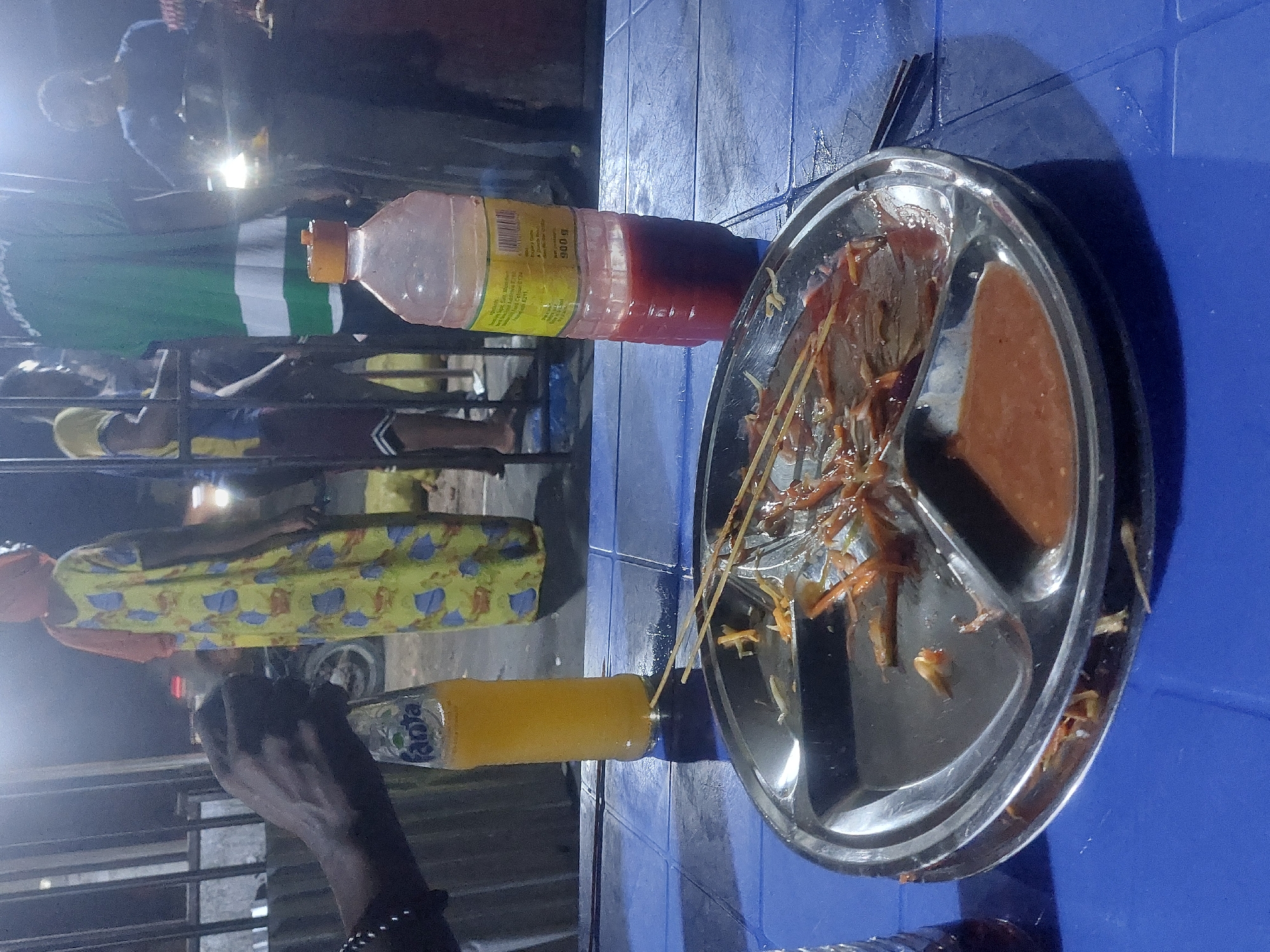 This photo captures a food stall scene at night in Dar es-Salaam, Tanzania. A blue plastic table is the focal point, hosting a steel plate divided into compartments. One compartment holds a reddish-brown sauce, while others contain remnants of skewered grilled meat (likely Mishkaki, a local specialty). Several wooden skewers lie on the plate, suggesting that food was recently served. Two bottles are positioned on the table: one contains a reddish liquid, and another is a bottle of Fanta. In the background, several people are visible in a casual street setting. One woman wears a colorful patterned dress, adding vibrancy to the scene. The lighting is ambient, from street lights or the food stall itself. The focus of the image is clearly on the local food.