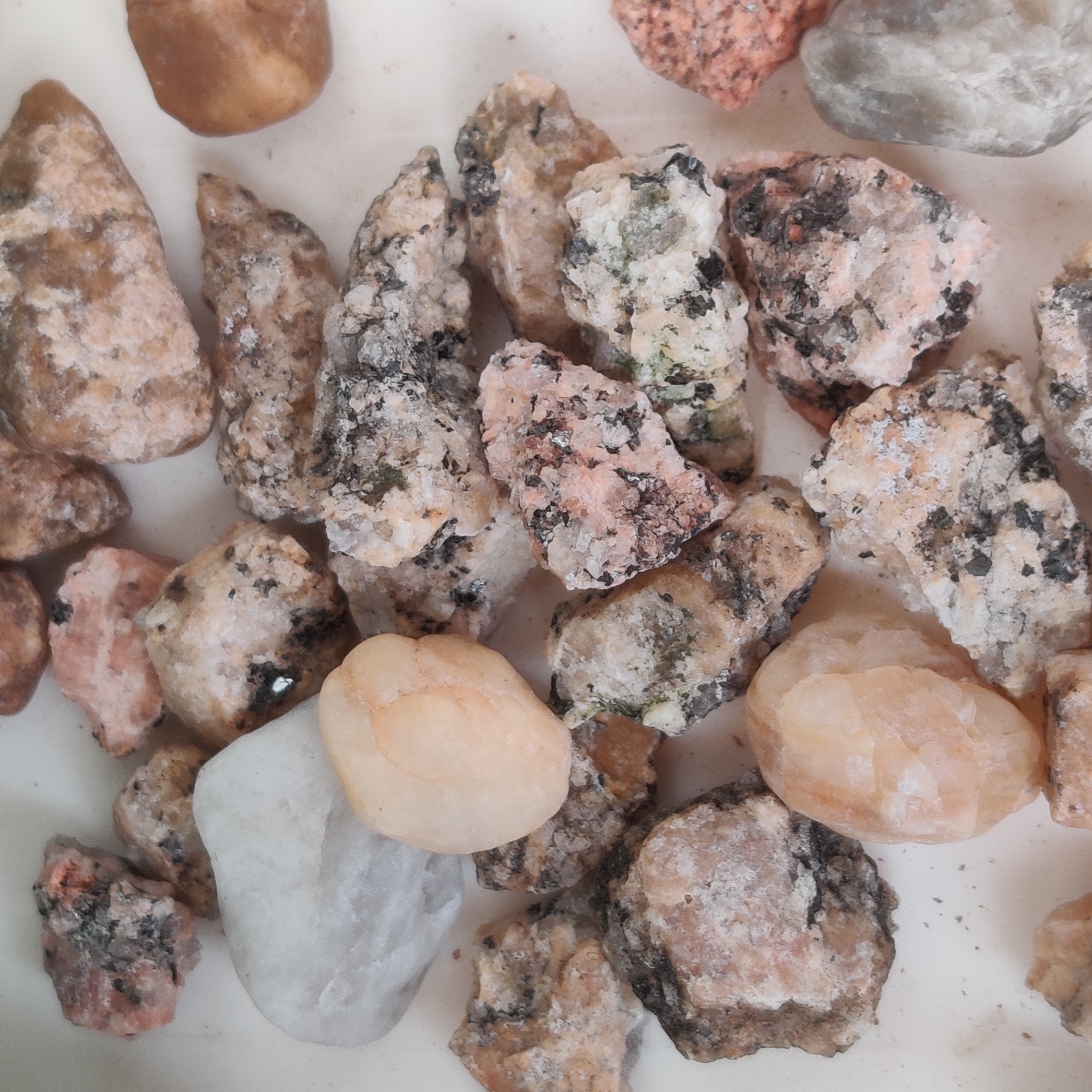 The image displays a collection of various rocks, primarily granite, scattered across a light-colored surface. The rocks exhibit a range of colors, including shades of pink, white, gray, and black, with some showing crystalline structures. The lighting is neutral, and the focus is sharp, providing a clear view of the textures and compositions of the rocks. There are no people or discernible actions taking place. The image does not contain any information or visual cues that would allow for the identification of a specific location in Yekaterinburg, Russia, or any other geographic area.