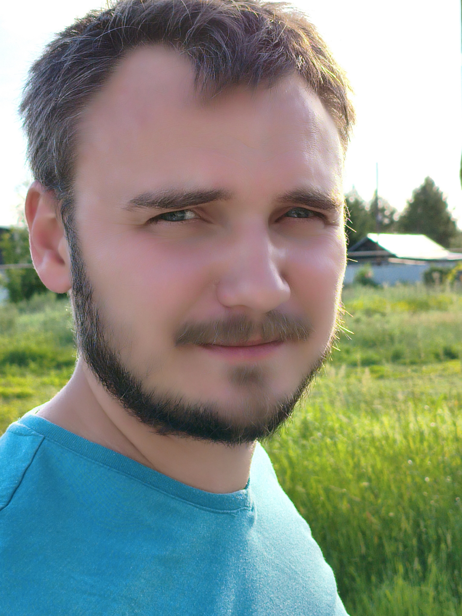The image is a close-up, outdoor, eye-level shot of a young Caucasian man. He has short, dark hair with graying at the temples and a short, dark beard and mustache. His eyes are light blue and he is looking slightly off-camera to his right. He is wearing a teal t-shirt. The background is out of focus and shows a grassy field with some trees and a small building, suggesting a rural or suburban setting. The lighting indicates it is daytime, possibly late afternoon due to the warm glow. No specific landmarks or indicators are present to pinpoint the location within Orenburg, Russia.