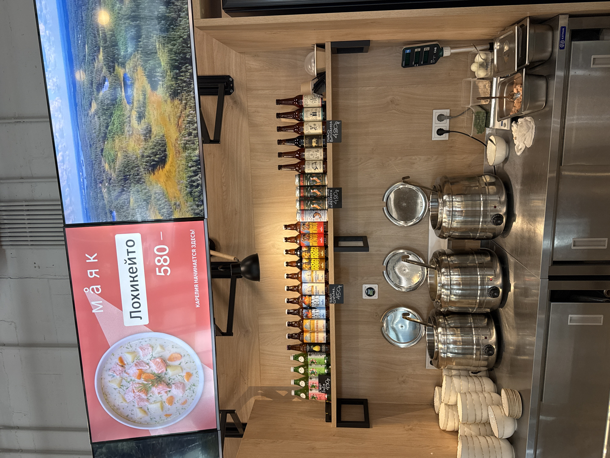 The image depicts an interior scene, likely a cafe or restaurant, with a strong focus on food and beverages. At the top, two large screens are mounted. The left screen displays a close-up of a creamy salmon soup with dill and what appear to be potato and carrot chunks, accompanied by Russian text: "МАЯК" (Mayak), "Лохикеито" (Lohikeitto), "580 -" and "КАРЕЛИЯ НАЧИНАЕТСЯ ЗДЕСЬ!" (Karelia begins here!). The right screen shows an aerial view of a forested landscape with numerous small lakes.
Below the screens, a wooden shelf is lined with an assortment of bottled beverages, predominantly beer, with some bottles featuring labels with Cyrillic text and prices indicated on small tags (e.g., "450р"). A black metal lamp with an articulated arm is positioned above this shelf.
Further down, a stainless steel counter features three large, silver soup tureens with ladles inserted. To the left of the tureens, stacks of white bowls and smaller dishes are arranged. To the right of the tureens, a scale is visible, along with several plastic containers holding food items, including what appears to be cooked salmon and hard-boiled eggs. A small white bowl and a crumpled white cloth are also present on the counter.
The environment appears clean and organized, suggesting a service area for customers. The presence of Russian text and the mention of "Karelia" strongly suggest the location is in Russia. Given the name "Lohikeitto" (a Finnish salmon soup) and the reference to Karelia, a region in northwestern Russia bordering Finland, it is highly probable that the establishment is located in or related to this geographical area. Lakhdenpokhya, Russia, is a city in the Republic of Karelia, making it a plausible location for this establishment.