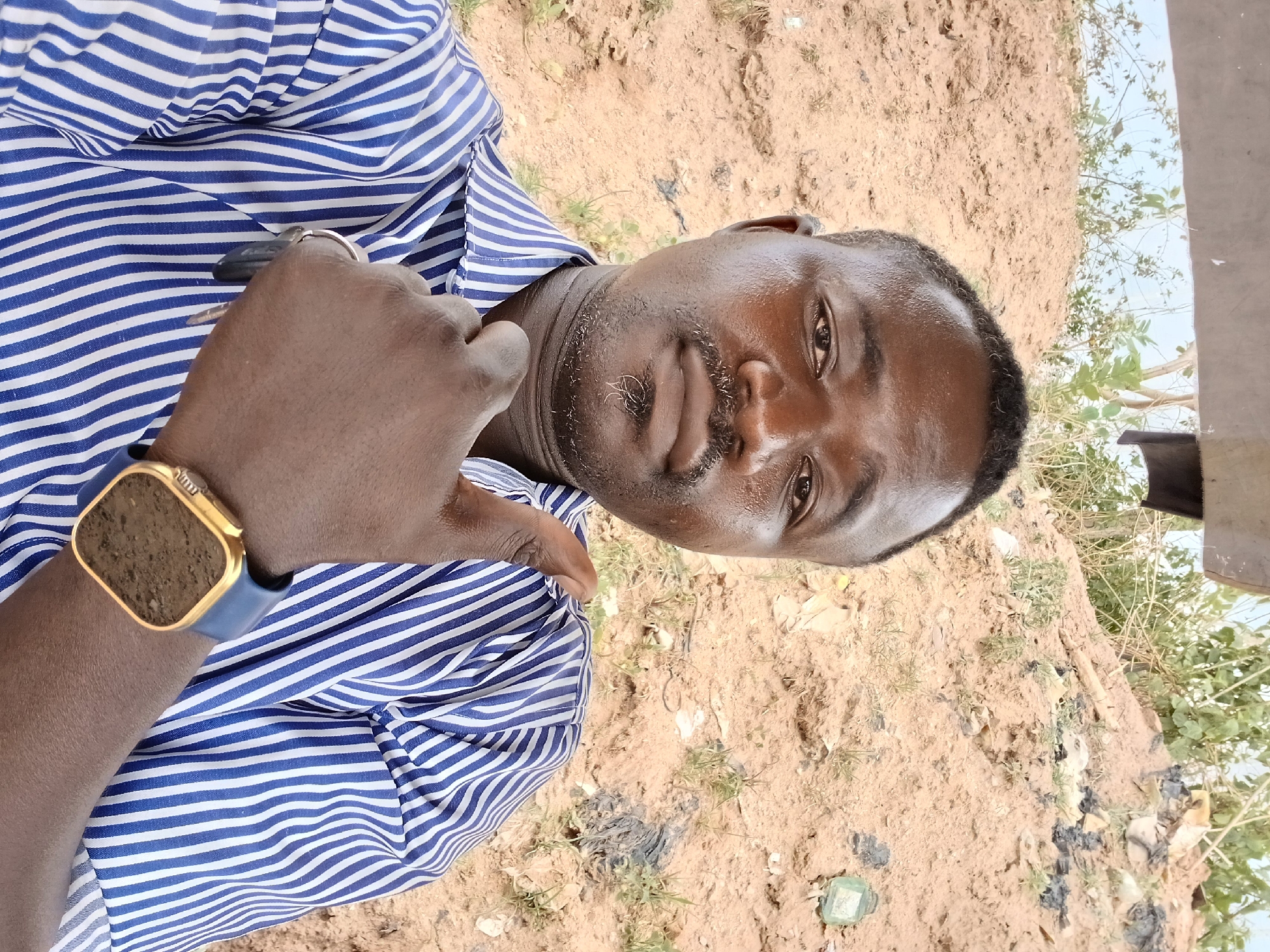 A man stands outside on dirt ground in Abuja, Nigeria. He has dark skin, short black hair, and a mustache. He wears a blue and white striped button-down shirt and a gold smartwatch with a blue band. He holds a black car key between his fingers. The background shows dirt ground with sparse greenery.