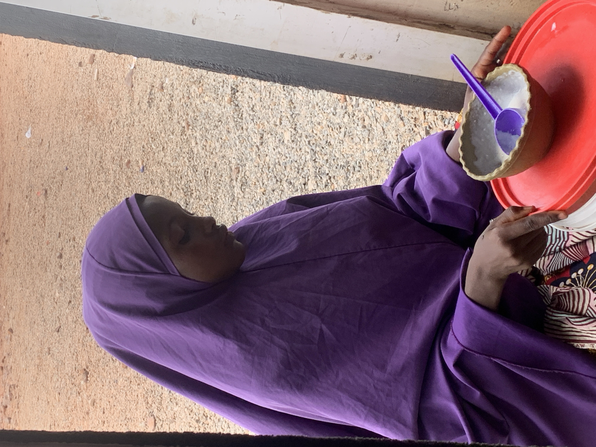 A woman wearing a purple hijab sits outdoors in Adankolo, Nigeria. She holds a red plate with a brown bowl containing a white substance and a purple spoon. The ground is gravel, and a gray stripe is visible at the top.
