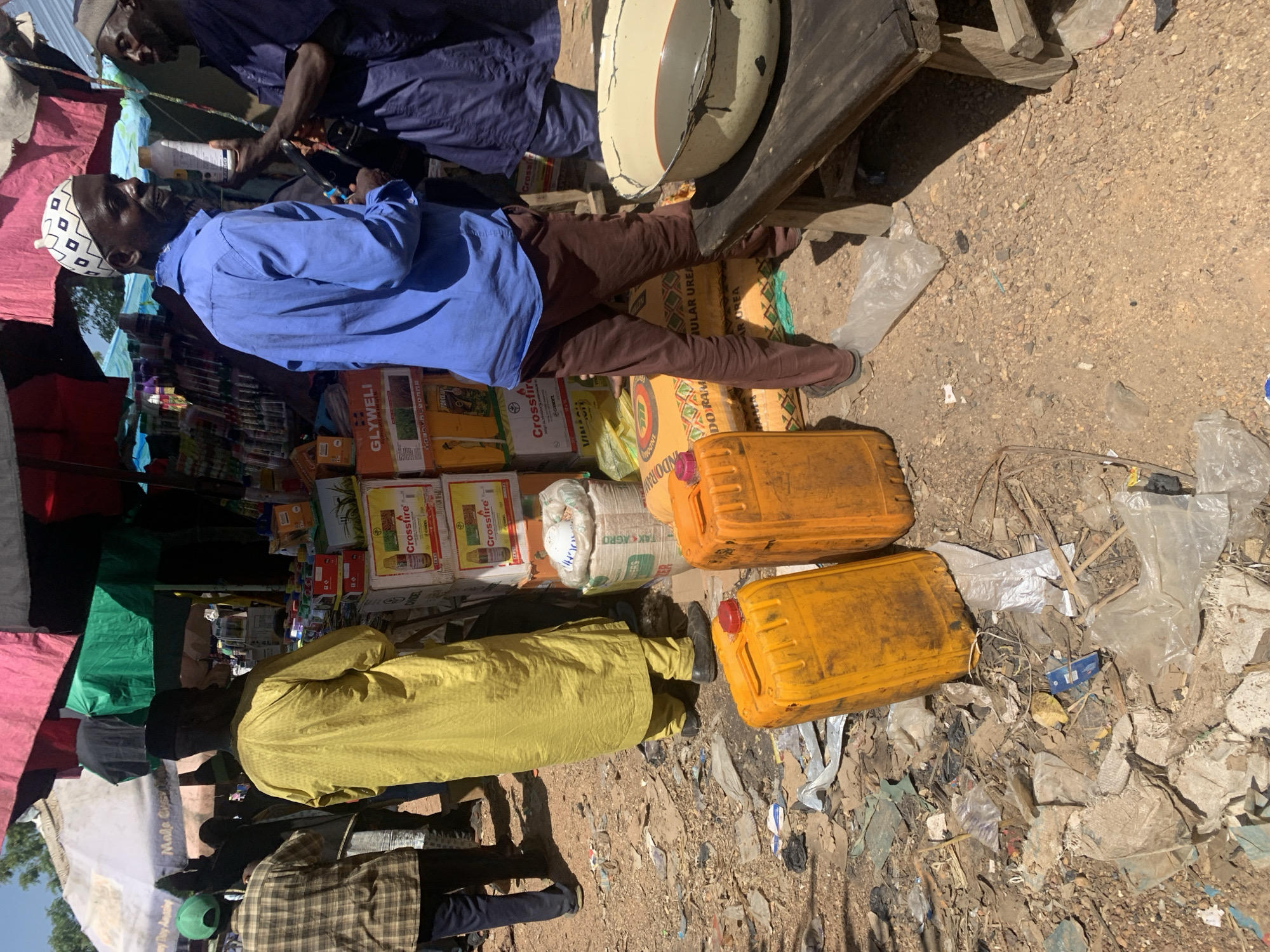 In Boi A, Nigeria, a bustling outdoor market scene unfolds under a canopy of colorful tarps. The ground is a mix of dirt and scattered debris, with plastic bags, wrappers, and other detritus.
In the foreground, two large, bright yellow plastic containers with red caps are prominently displayed. To their right, a large, rustic wooden table holds a metal basin filled with a dark liquid.
Several individuals are present, engaged in various activities. A man in a blue long-sleeved shirt and brown pants, wearing a distinctive white and black patterned cap, is holding a white bottle and appears to be in conversation with another man dressed in a dark blue robe, who is also holding a white bottle. Behind them, another man in a yellow robe stands with his back to the camera. Further back, other figures are visible, including a person in a plaid shirt and green cap.
The market stalls are laden with goods, including stacks of boxed items, suggesting a variety of products for sale. The overall atmosphere is one of commerce and daily life in a Nigerian market.