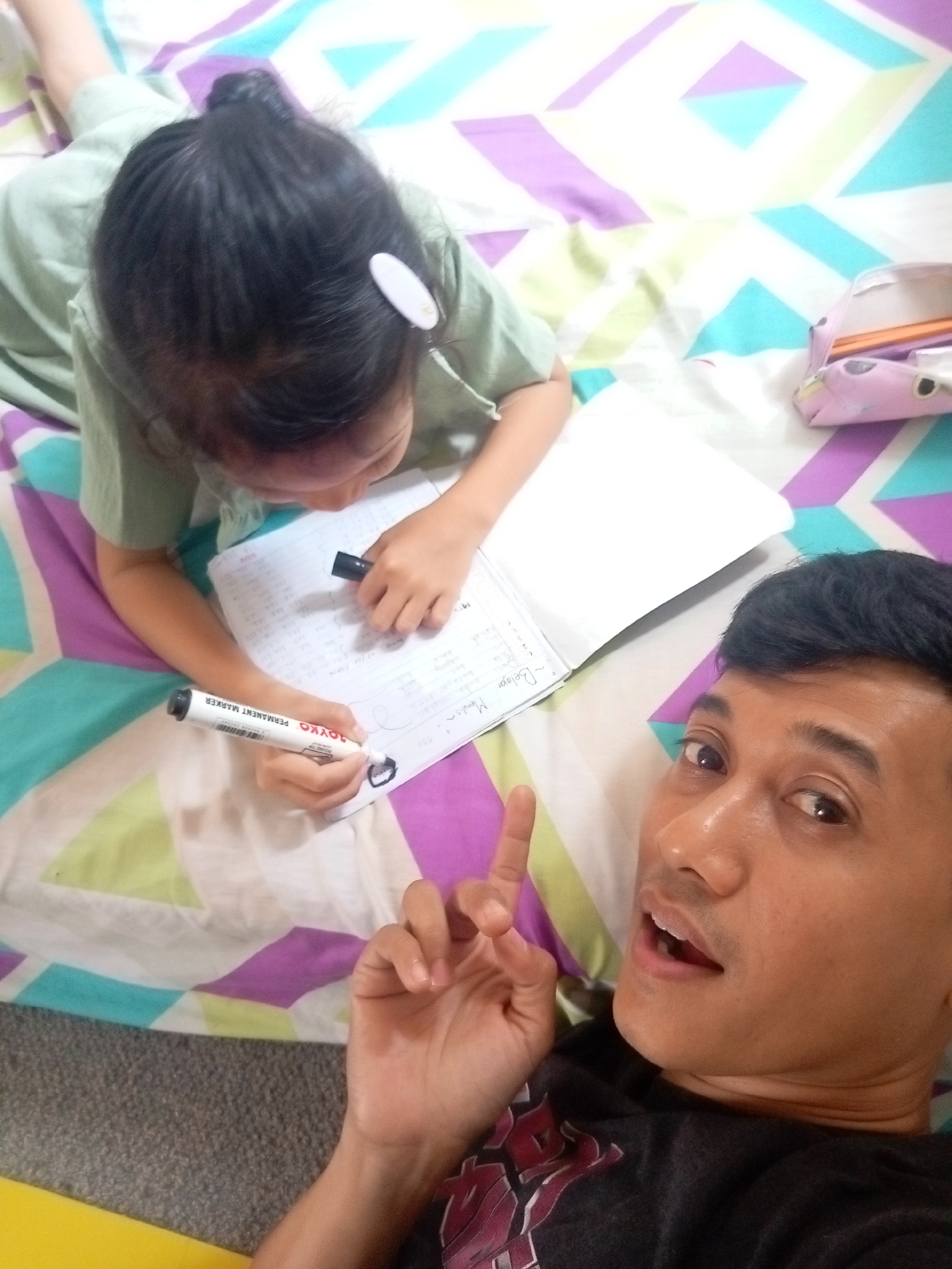 A man and a young girl are lying on a colorful geometric patterned bed. The man, positioned in the foreground on the right, is looking at the camera with his mouth open, and his right hand is raised in a peace sign. He is wearing a dark t-shirt with a red and black graphic design. The young girl, on the left, is leaning over an open notebook, writing with a black marker. She is wearing a light green t-shirt and has her hair pulled up in a bun, secured with a white clip. A white marker with black lettering, "JOYKO PERMANENT MARKER," is in her hand. Next to the notebook is a pink pencil case. The background is a bed with a vibrant, abstract geometric pattern of purple, green, blue, and white shapes. The lighting suggests an indoor environment, possibly a bedroom.