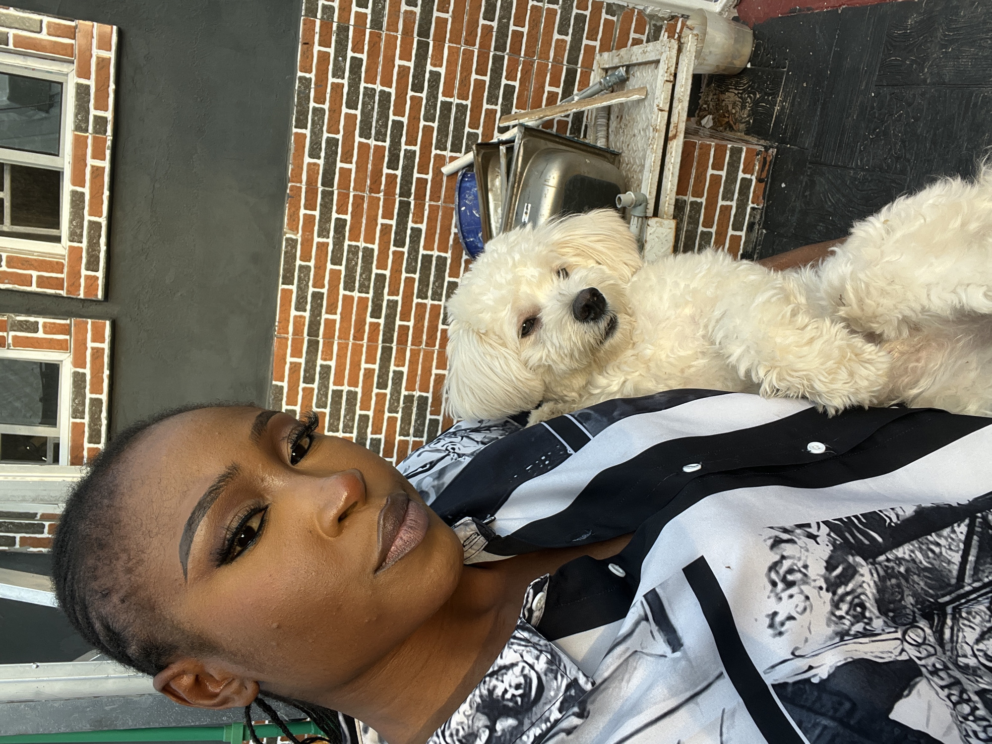 A woman with dark skin and braided hair is holding a small, fluffy white dog. She is wearing a black and white patterned shirt. The dog is looking towards the camera. In the background, there is a brick wall with windows and some metal structures, possibly part of a construction site or a utility area. The setting appears to be outdoors.