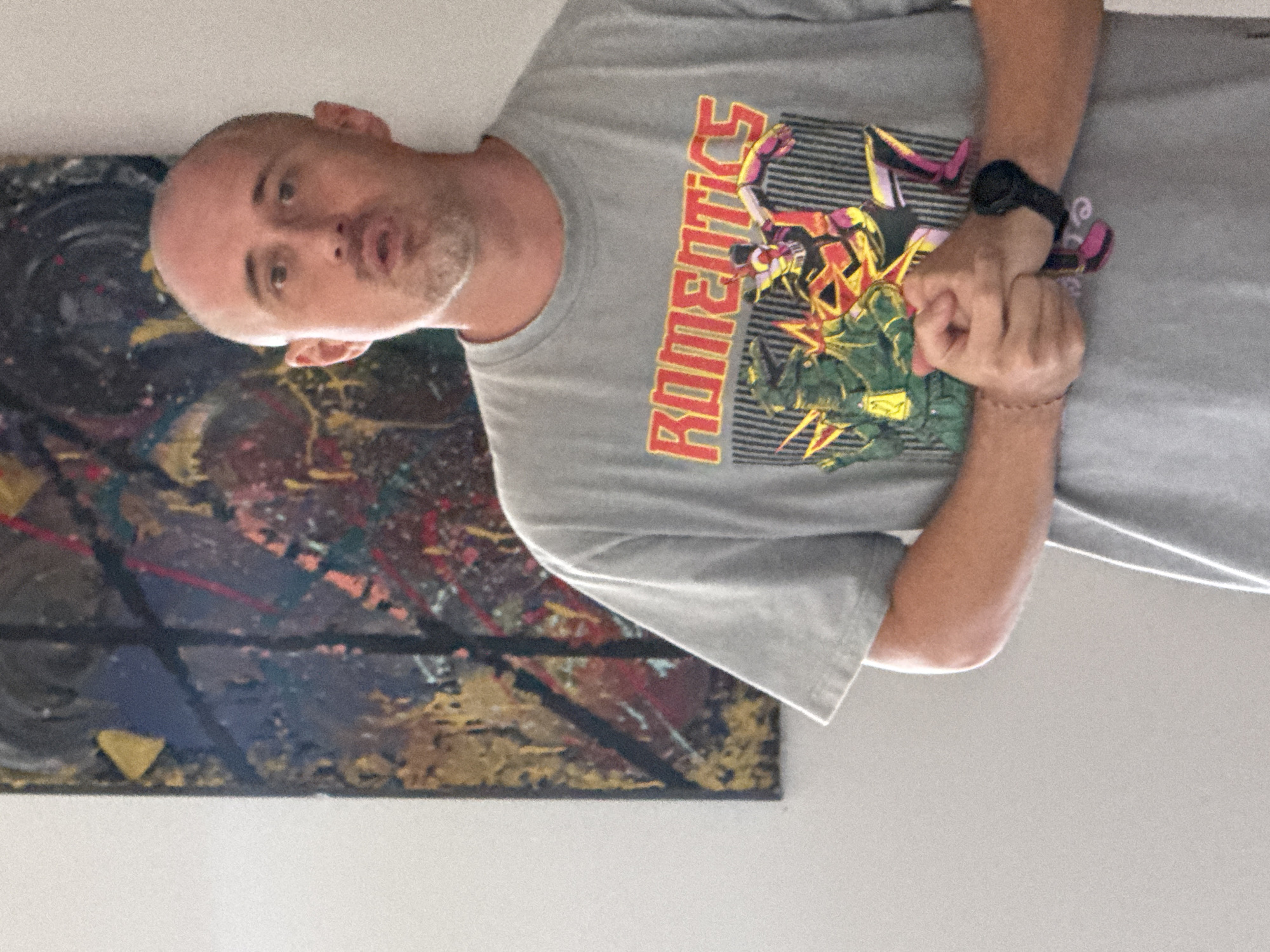 The image displays a man in an indoor setting, identified as Denpasar, Indonesia. He appears to be middle-aged with a bald head and light stubble on his chin. He is wearing a light grey short-sleeved t-shirt that features a prominent graphic. The graphic includes the word "ROMENTICS" in red capital letters with a yellow outline, positioned above two stylized robot figures engaged in an action pose, one in pink/purple and yellow, the other in green and yellow, set against horizontal grey stripes.
The man's hands are clasped together at his waist. He wears a black digital watch on his left wrist and a thin, dark-colored bracelet on his right wrist. His mouth is slightly open, suggesting he might be speaking or about to speak, and he looks towards the viewer. Behind him, on a light-colored wall, hangs a large abstract painting. This artwork features a dark background with dynamic splatters and strokes of red, gold, yellow, and other colors. The overall scene captures a casual moment in what appears to be a home or studio environment. The image itself is rotated ninety degrees clockwise.