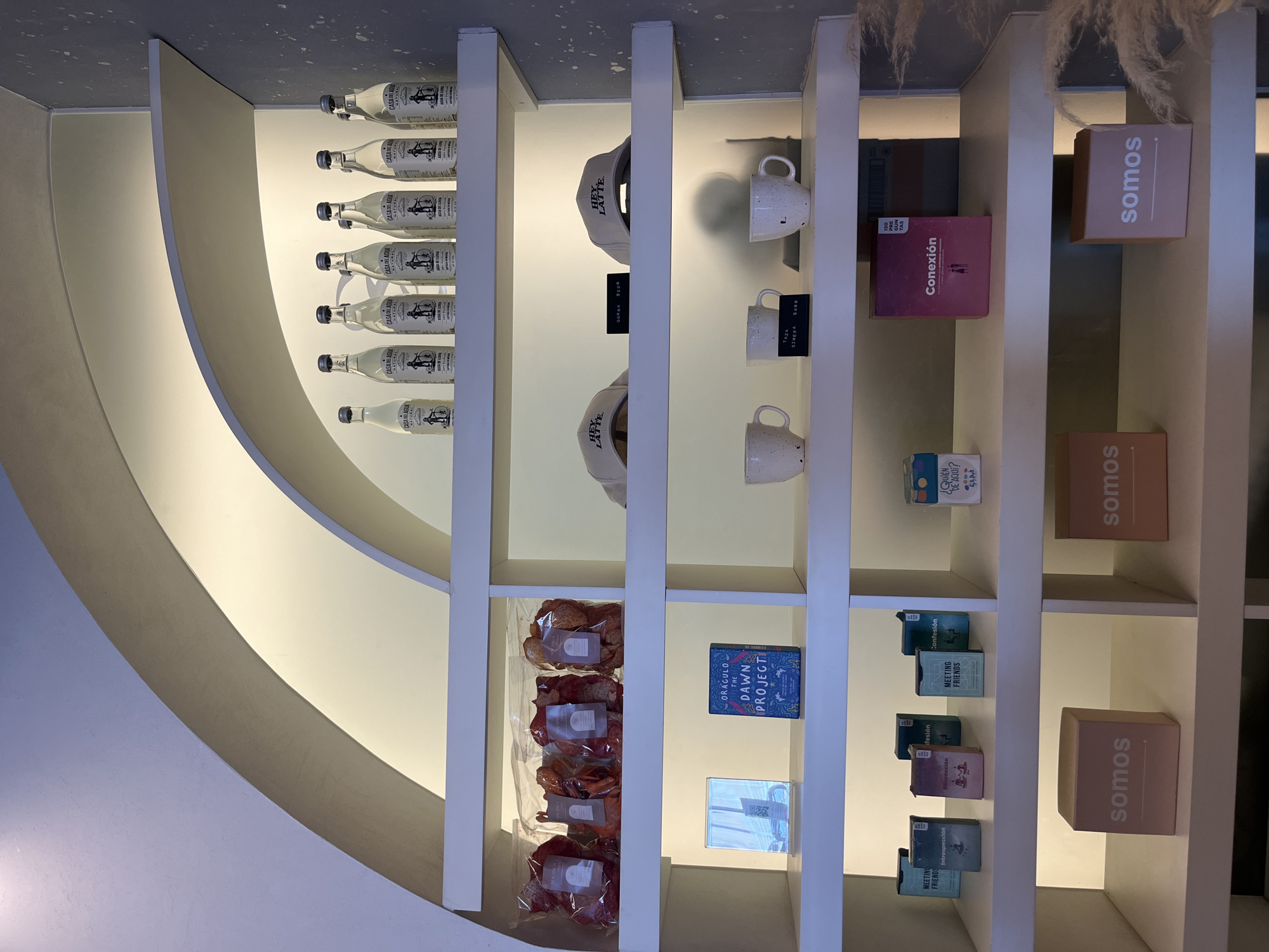 The image presents a display shelf within a commercial establishment, likely a store or boutique, in San Bernardino Tlaxcalancingo, Mexico.
The shelves are white and arranged vertically, displaying various items:
- Bottles of "CASA DEL AGU" (mineral water) along the far left.
- "HEY LATTE" baseball caps.
- "TAZA SIMERA" cups.
- Books labeled "Conexión", "ORAGULO THE DAWN PROJECT", "MEETING FRIENDS", and "¿QUIÉN DE AQUI?".
- "somos" branded boxes.
- Packaged food items resembling chips.
The composition is a shelf display with warm lighting.