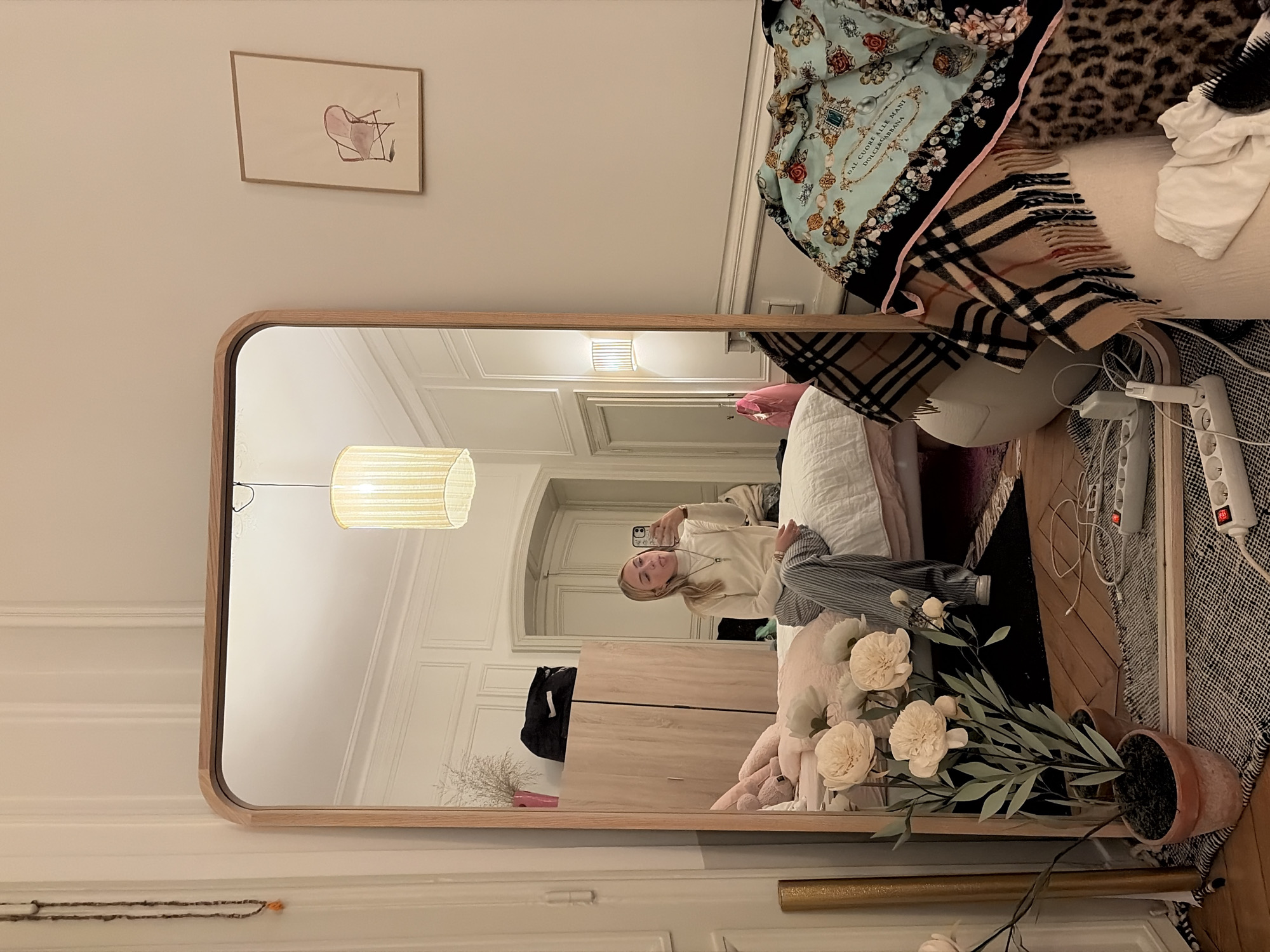 This image depicts a bedroom interior within a Haussmannian apartment in Paris, France, characterized by its high ceilings, ornate wall paneling, and tall doorways. The scene is primarily captured through a large, rectangular, light wood-framed full-length mirror leaning against a wall.
Reflected in the mirror, a young Caucasian female with blonde hair, wearing a cream turtleneck and striped trousers, is seated on a bed. She holds a smartphone, capturing a selfie, with her tongue playfully extended. The bed has white bedding, and a pink plush toy is visible. Behind her, a light-toned wooden wardrobe holds a pink-potted plant with dried grass. A minimalist ceiling lamp with a fabric shade illuminates the room. An arched doorway and a wall sconce are visible in the background.
The foreground, containing the mirror, features a wooden floor partially covered by a black and white patterned rug. A potted plant with large artificial white peonies stands to the left of the mirror on the rug. To the right of the mirror, a white power strip with multiple outlets and a red switch, along with several cables, rests on the rug. A collection of items is piled on a white, rounded stool-like object: a beige plaid scarf, a light blue patterned scarf with the text "DAL CUORE ALLA MANI DOLCE&GABBANA," a leopard print blanket, a white fabric item, and a black hairbrush. On the wall adjacent to the mirror's left side, a long beaded necklace with an orange accent hangs. Above the mirror's upper right corner, a framed minimalist artwork of a pink armchair is displayed.