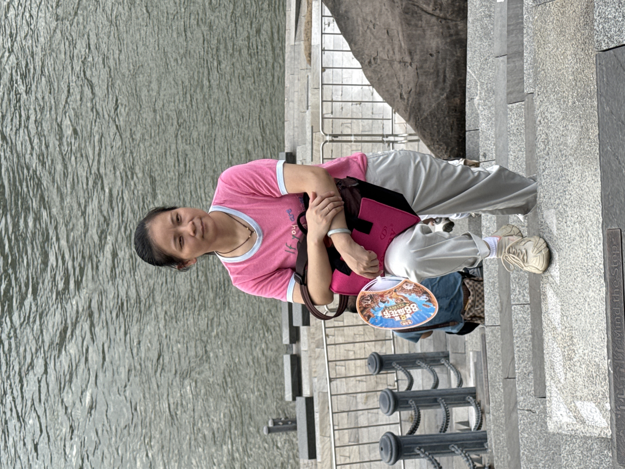 A woman stands on a stone staircase next to a body of water, possibly the Minjiang River, in Nan'an District, China. She is wearing a pink t-shirt with a light blue trim, light-colored pants, and beige sneakers. She is holding a large pink tote bag and a circular fan with an image on it. Her left arm is crossed over her chest, and she is looking to her right. Behind her, a metal fence separates the staircase from the water, and other people are visible further down the stairs. A large, dark rock formation is to her right. The staircase has an inscription at the bottom that reads "Mind the Step".