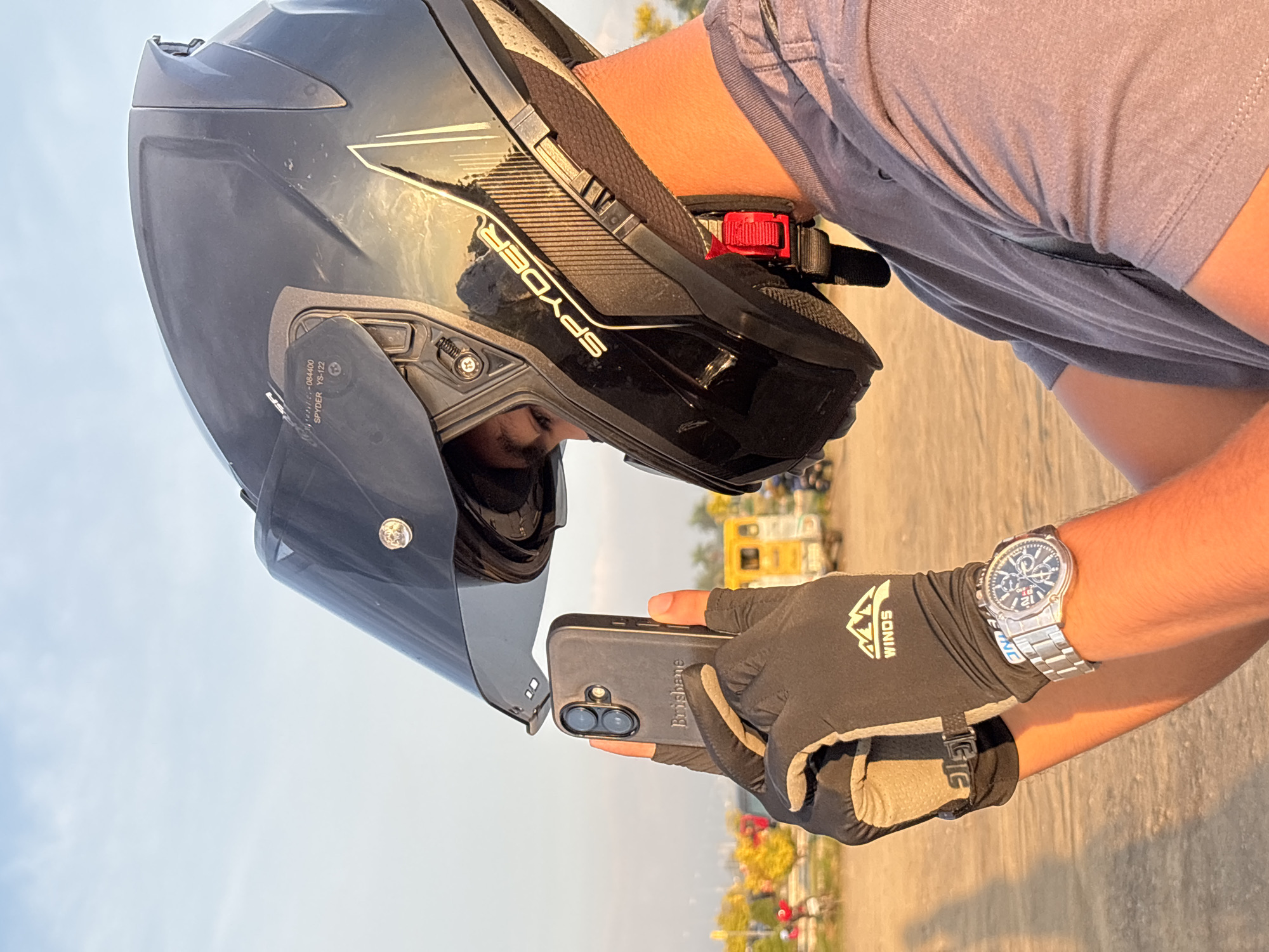 A person wearing a black Spyder motorcycle helmet and a grey t-shirt is holding a black smartphone with a grey case. The person is also wearing black riding gloves with a "WINDS" logo and a silver wristwatch. The background appears to be an outdoor setting with a dirt ground and a yellow vehicle in the distance. The sky is clear and blue. The scene takes place at the coastal road near the San Juanico Strait Bridge in Davao City, Philippines. The person is in the process of taking a photo of themselves or the surroundings with their smartphone.