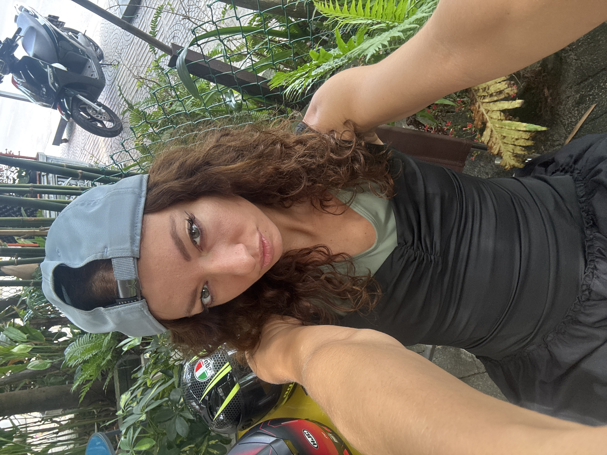 A young woman with curly brown hair is taking a selfie in an outdoor setting in Đà Nẵng, Vietnam. She's wearing a grey baseball cap and a black top. Visible is a portion of a yellow and black motorcycle helmet, as well as a motorcycle parked in the background. Green foliage, including ferns, surrounds the area, and a wire fence is visible behind her. The ground appears to be paved.