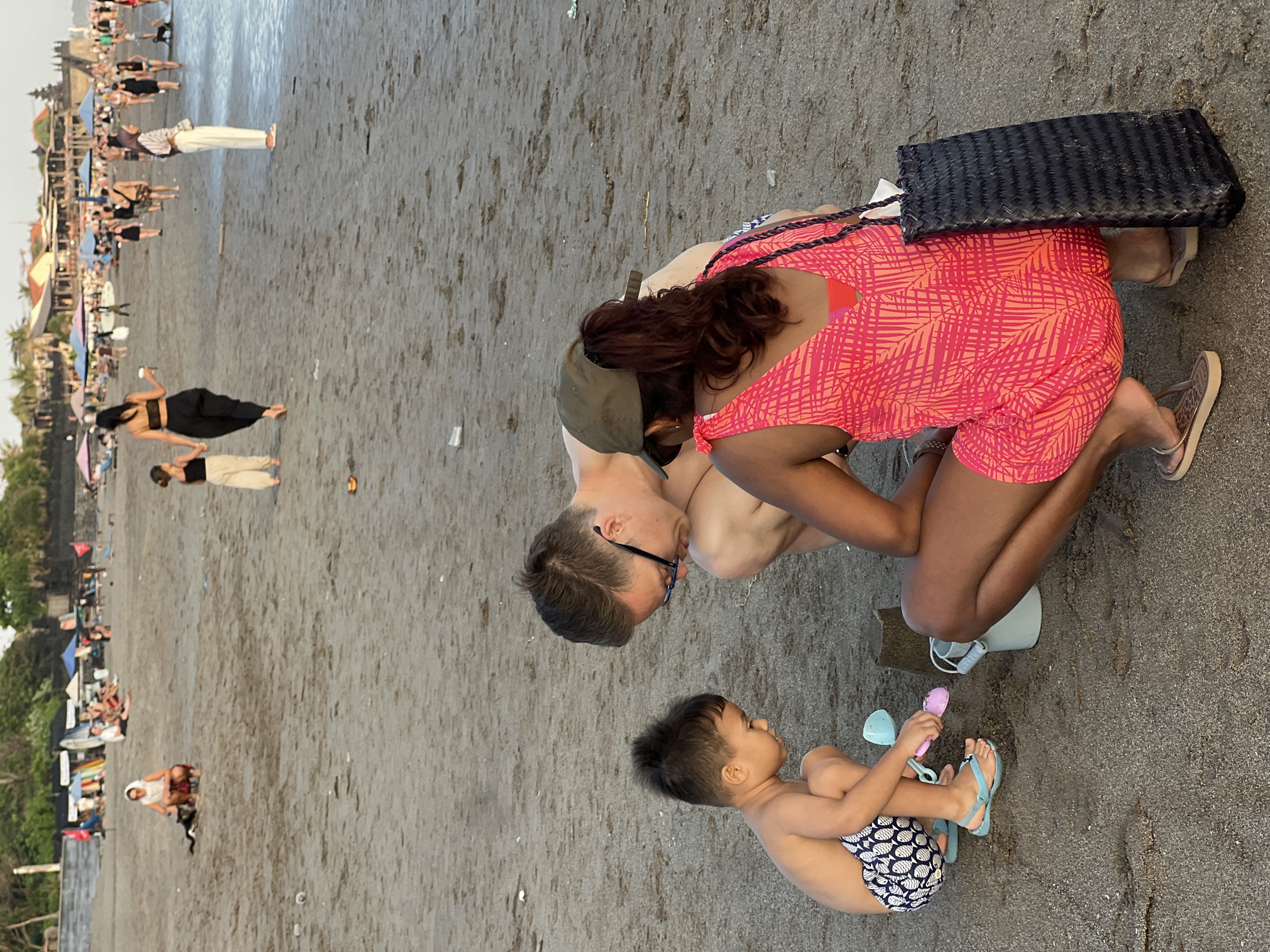 In North Kuta, Indonesia, at sunset on a sandy beach, a family is engaged in a playful activity with sand toys. A young boy, wearing patterned swim trunks and blue flip-flops, is intently focused on building a sandcastle with a pink shovel and a bucket. He is crouched down, his body angled towards the sand. Beside him, a man with short brown hair and glasses is also crouched, observing the boy's efforts. Partially visible in the foreground, a woman with dark hair and a green cap is kneeling, wearing a vibrant pink and orange leaf-patterned dress. She has a woven black bag resting beside her. The background of the image reveals other beachgoers enjoying the late afternoon light, with a line of buildings and palm trees visible further inland. The sand is a light brown, with footprints and scattered debris indicating recent activity. The ocean is not clearly visible, but the overall ambiance suggests a serene beach scene.
