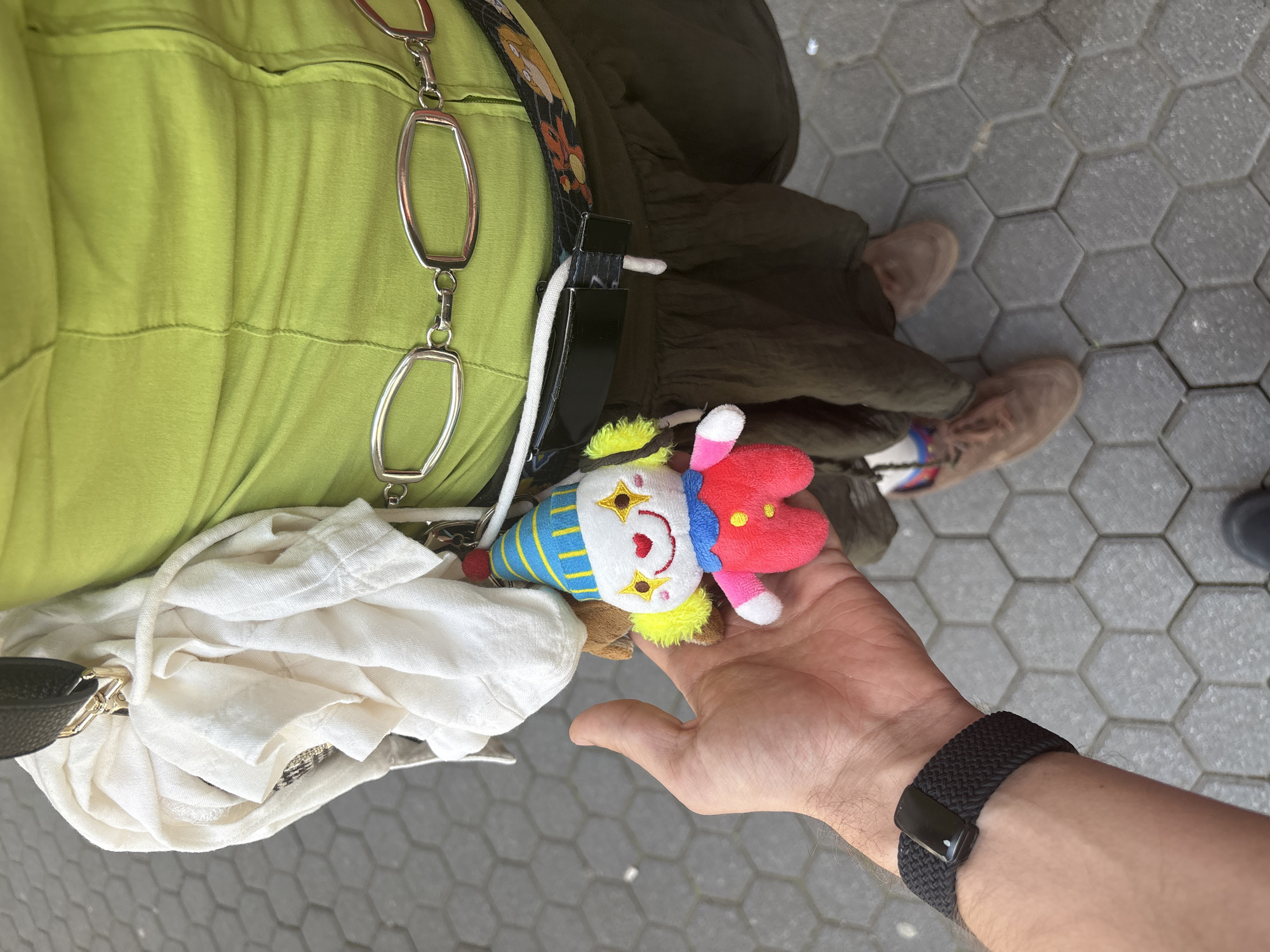 A first-person, overhead shot shows a person holding a small clown doll in their right hand. The doll has a blue and white striped hat, a white face with yellow star details, yellow hair, and a red headpiece. The person is wearing a black watch band on their wrist. In the background, another person stands, wearing a light green dress or top with a silver chain detail, an olive green skirt, and tan shoes. A white cloth bag is draped on the left side of the frame. The ground is paved with hexagonal grey pavers. This scene is located in Sofia, Bulgaria.