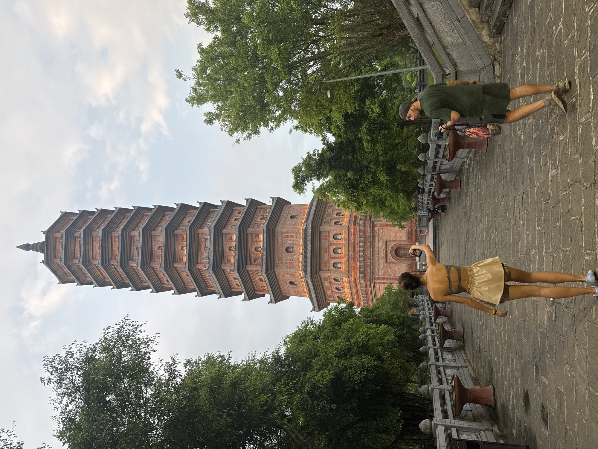 A medium-shot, eye-level, outdoor, daytime, full-color photograph shows a brick pagoda in Tây Hoa Lư Ward, Vietnam, with two people in the foreground. The pagoda is tall and multi-tiered, constructed of red brick, with decorative features and illuminated windows. Lush green trees flank the pagoda and the pathway leading towards it. The pathway is paved with stone slabs and lined with decorative balustrades and planters.
In the foreground, a woman is walking away from the camera, turned slightly to her left, holding up a smartphone to take a photo. She is wearing a strapless grey top and a light-colored skirt. To her right, a man is walking away from the camera, looking over his left shoulder. He is wearing a dark green t-shirt, dark shorts, a baseball cap, and carrying a patterned bag. The sky is overcast with some clouds.