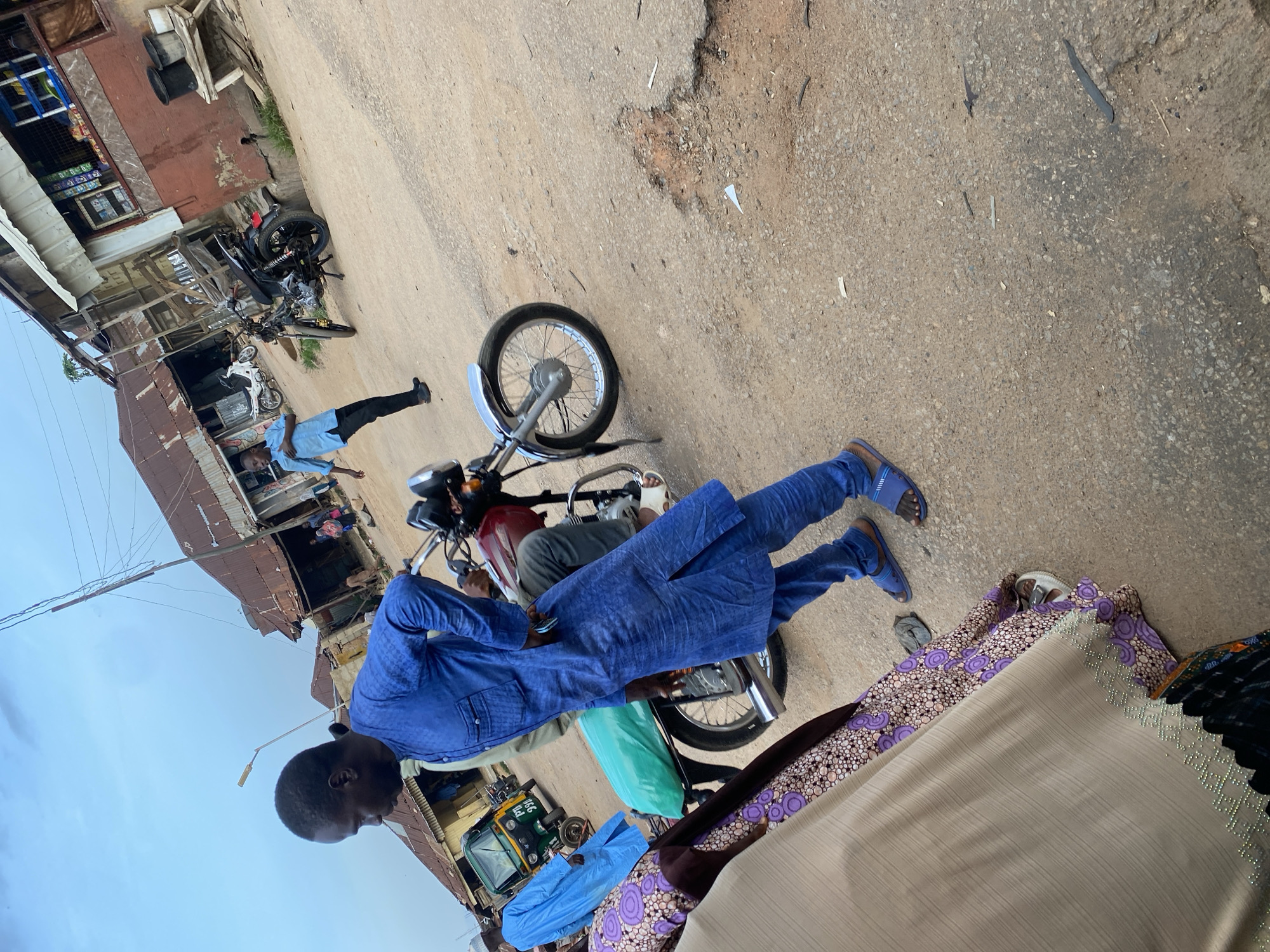 The image depicts a street scene in Jos, Nigeria. A man dressed in a blue traditional outfit stands by a red motorcycle, which is parked on a dirt road. The road is uneven and shows signs of wear and tear, with a large pothole visible in the foreground. Behind the man and motorcycle, other individuals and vehicles are present, including a green auto-rickshaw and another motorcycle. The background features buildings with corrugated iron roofs, typical of many urban areas in Nigeria, some appearing weathered and in disrepair. The sky is overcast, suggesting it might be a cloudy day. The overall environment suggests a bustling, possibly commercial area with a mix of everyday life and transportation.